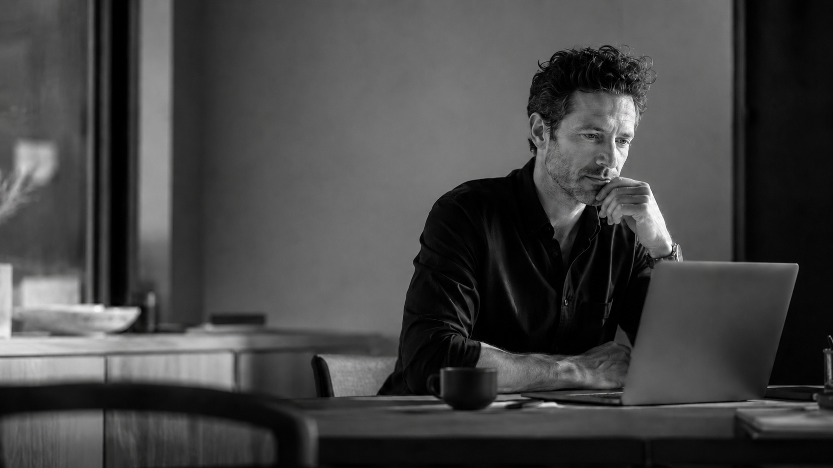 A man with curly hair and a beard sitting at a desk, looking at a laptop with a thoughtful expression, in a black and white photograph.