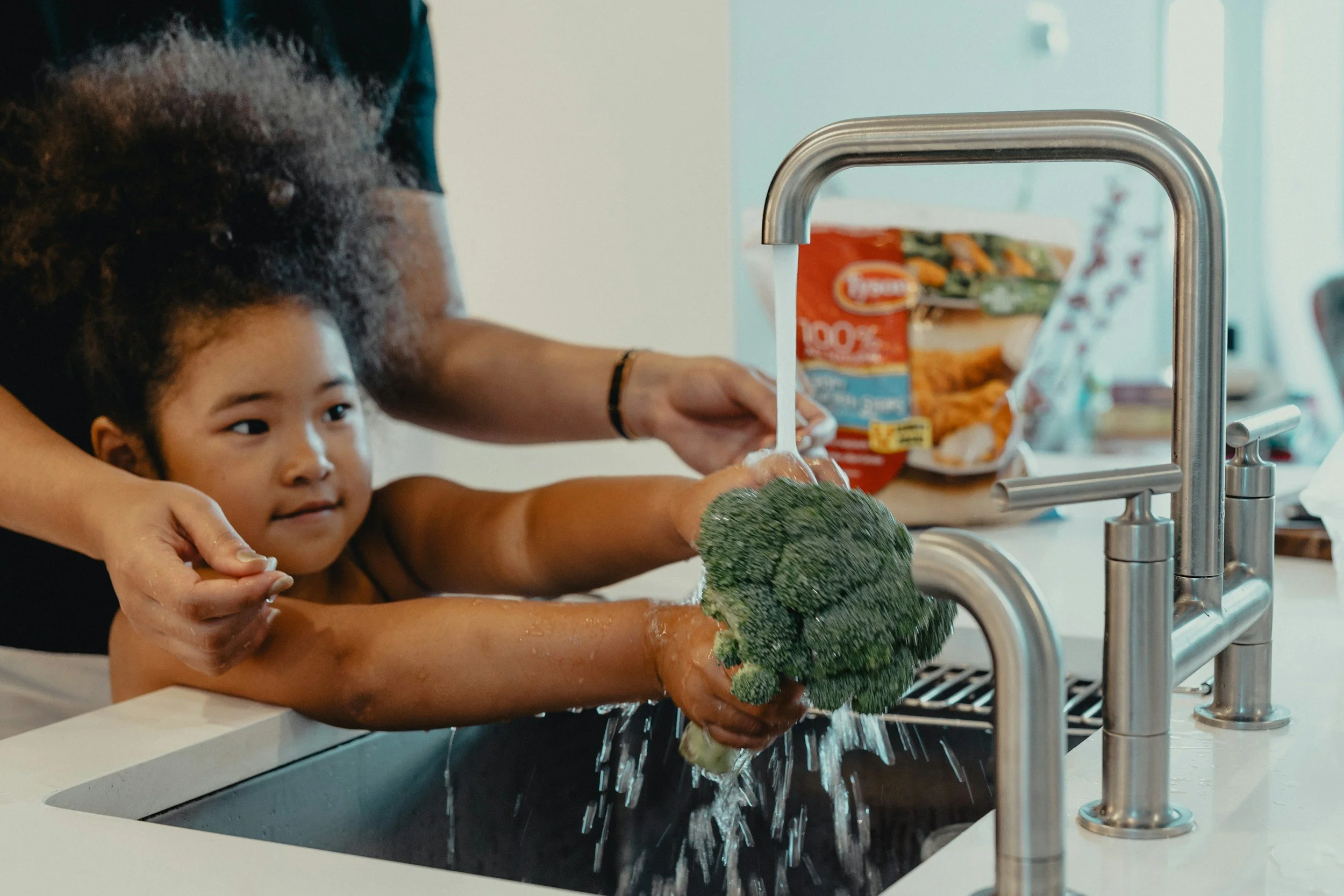 A young girl is washing a large broccoli under a kitchen sink faucet, assisted by an adult. The girl looks focused while water runs over the broccoli, with various food items and packaging visible in the background.
