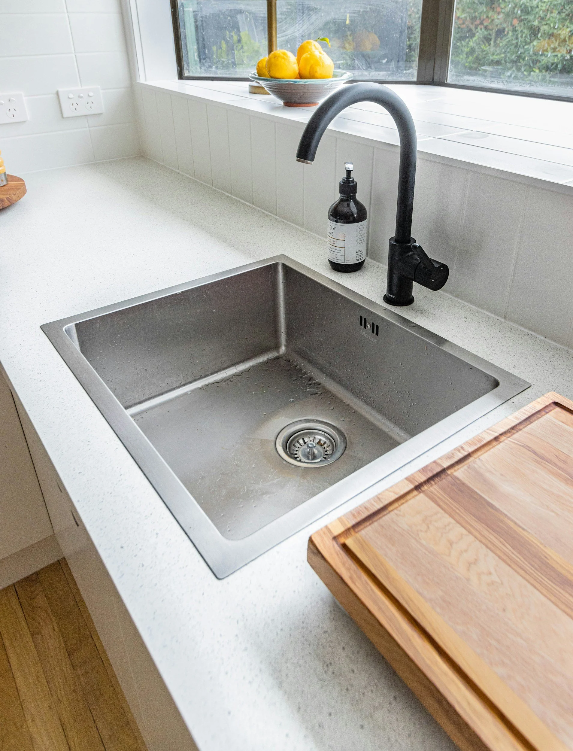 Kitchen sink with a black faucet, a soap dispenser, a wooden cutting board on the counter, lemons on a window sill, and an electrical outlet on a white tiled wall.