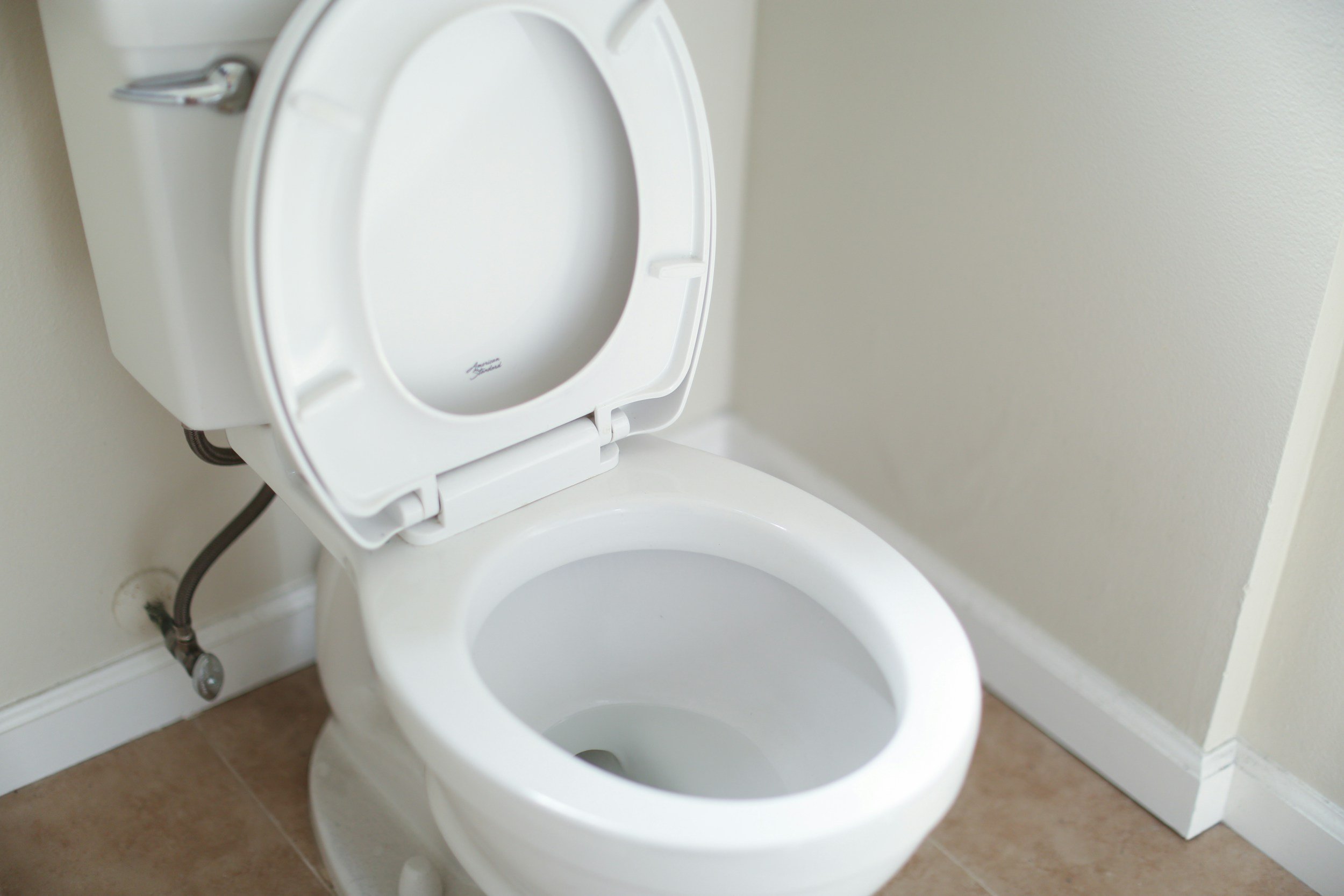 A white toilet with its lid open in a bathroom with beige floor tiles and cream-colored walls.