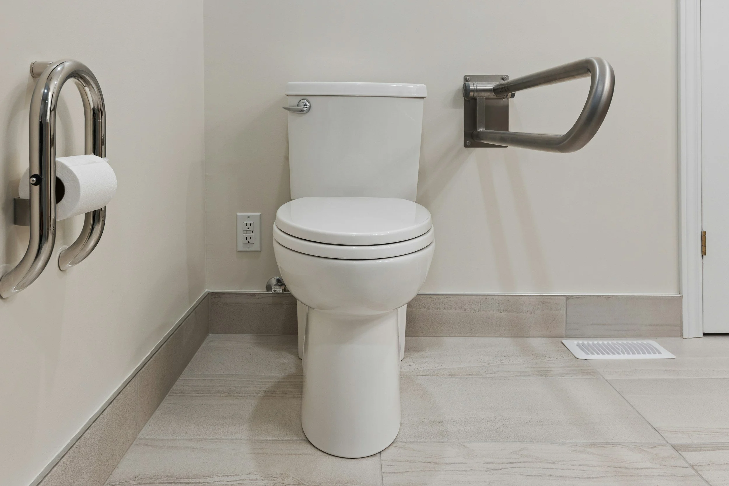 Toilet with grab bar on the wall to the right, wall-mounted toilet paper holder on the left, and a vent on the floor near the door.