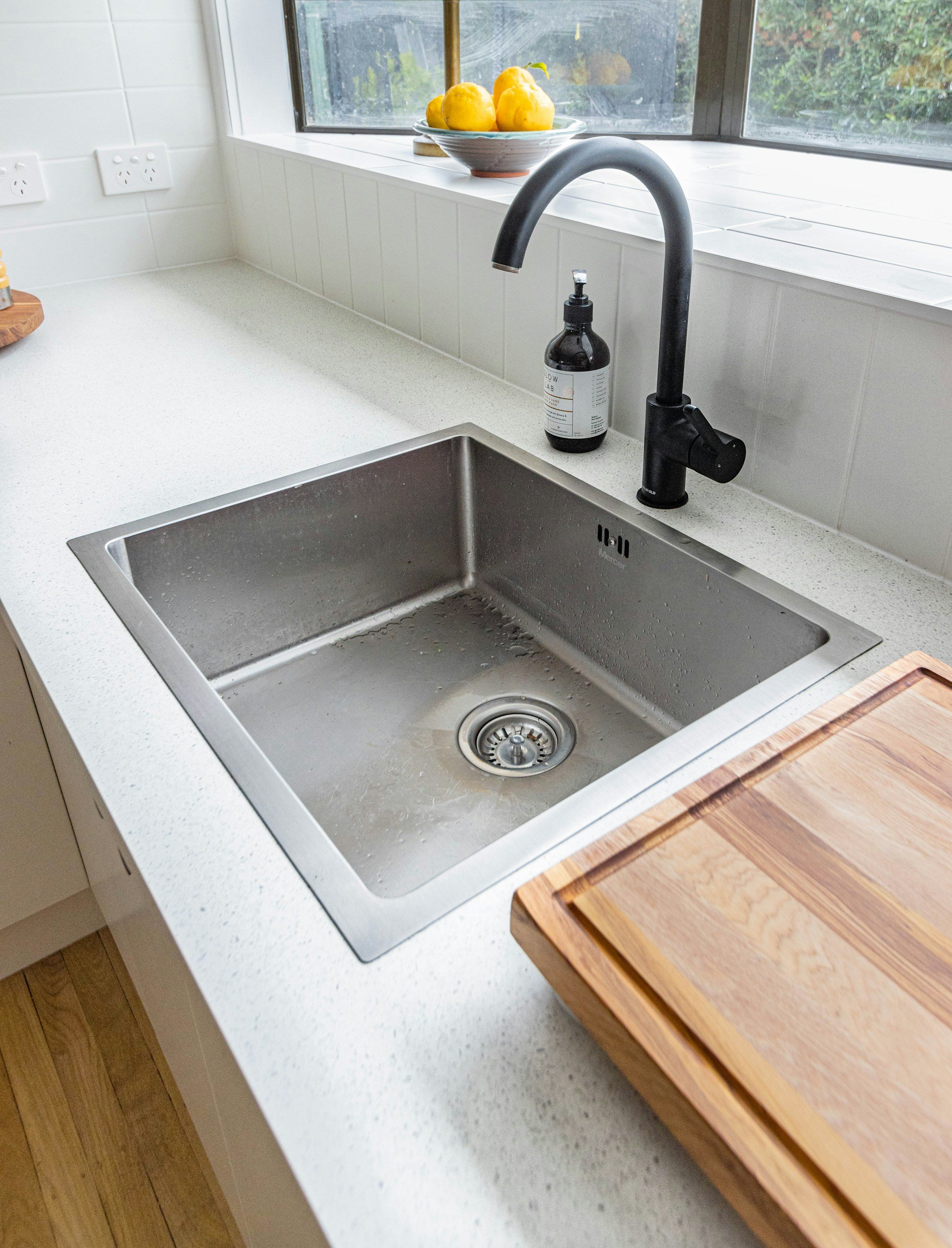 A kitchen sink with a black faucet, a soap dispenser, a wooden cutting board, and a window with a bowl of yellow lemons on the windowsill.