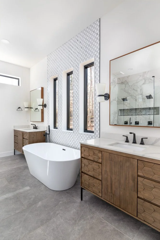 Modern bathroom with a freestanding bathtub, wooden vanities with black faucets, marble accents, and three tall narrow windows