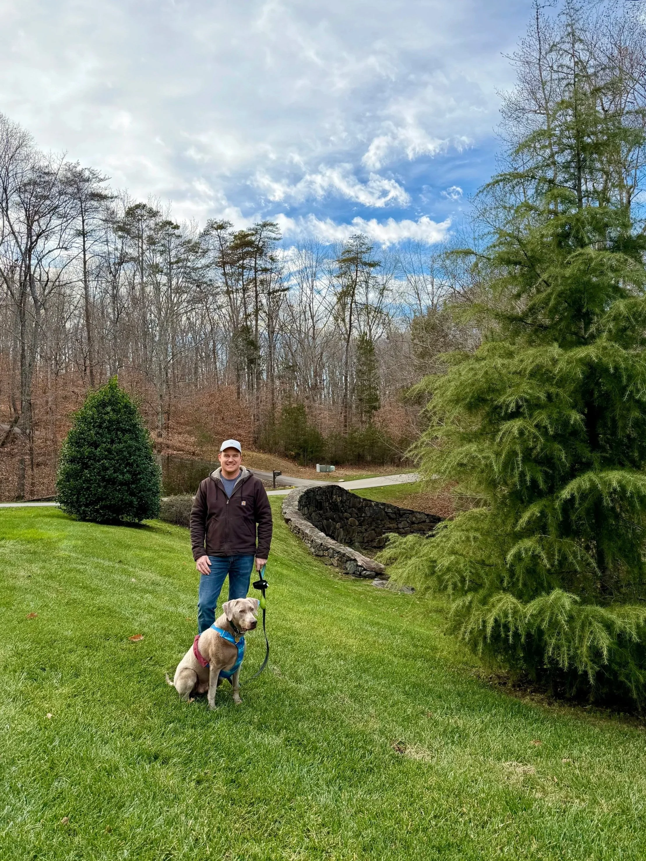 A man stands outdoors on a lush green lawn holding a leashed dog. The man is dressed casually in a jacket, jeans, and a baseball cap. The dog, a large, light-colored breed, is sitting on the grass next to him, wearing a harness. In the background, there are trees, a small stone wall, a dirt path, and a partly cloudy sky.
