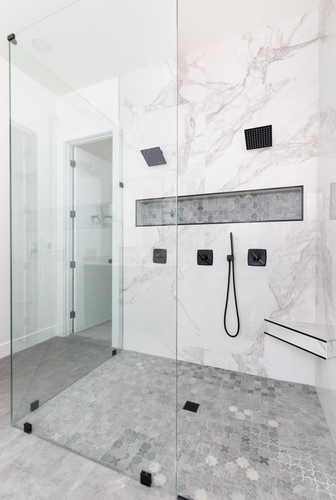 Modern walk-in shower with white marble walls, gray patterned floor tiles, black square controls, a handheld showerhead, two wall-mounted rainfall showerheads, a horizontal niche with decorative tile, and a glass enclosure.