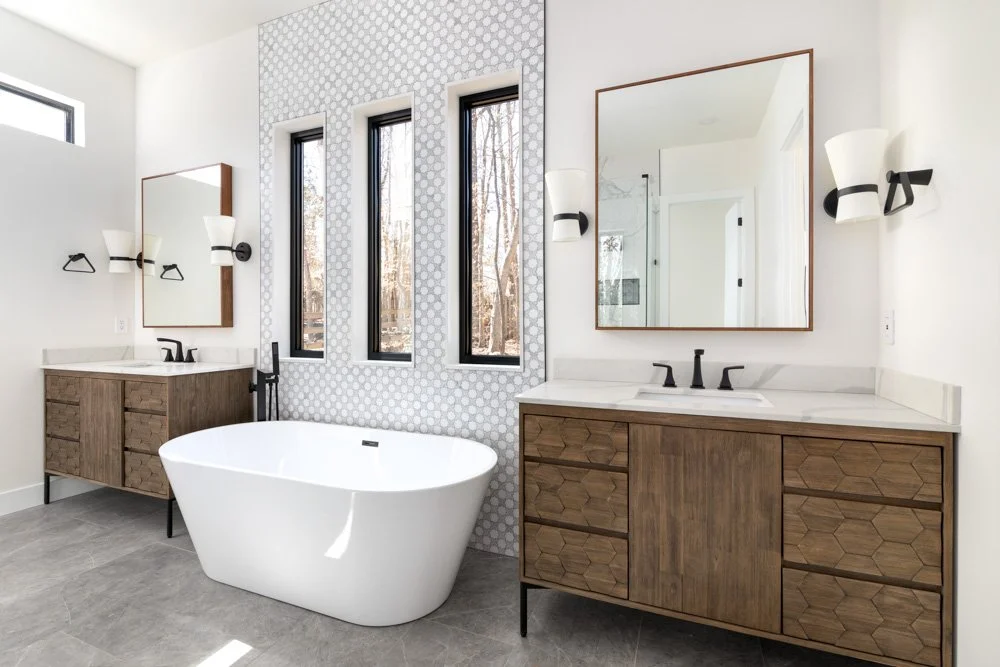Modern bathroom with two wooden vanities, large mirrors, a freestanding white bathtub, and three narrow windows with a view of trees outside.
