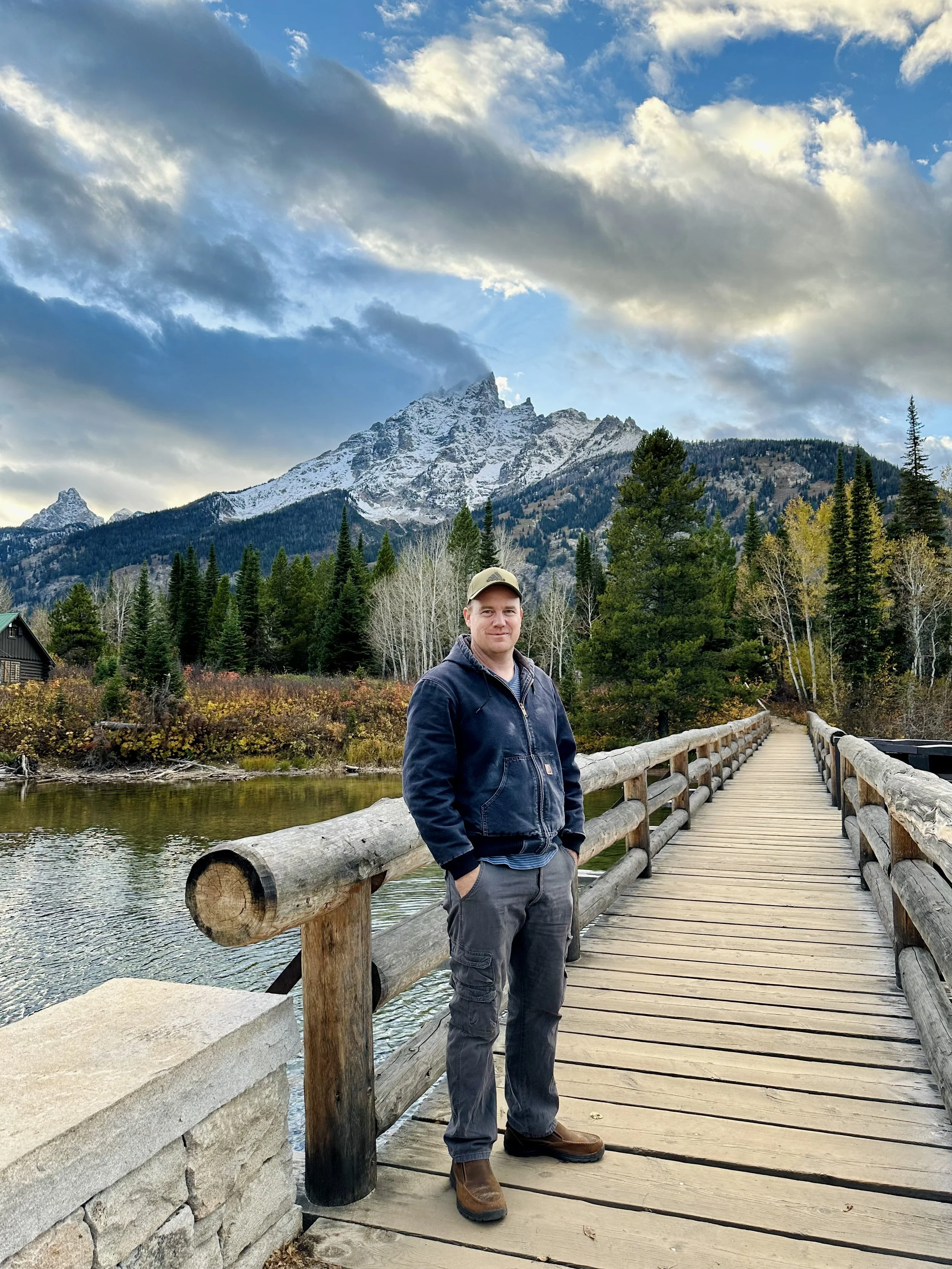 A man standing on a wooden bridge with a mountain and forest in the background during autumn.