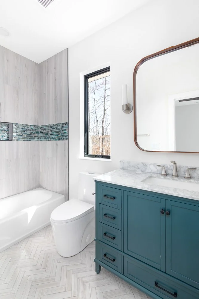 Modern bathroom with teal vanity, marble countertop, large mirror, white toilet, bathtub, mosaic tile accent wall, and window with trees outside.