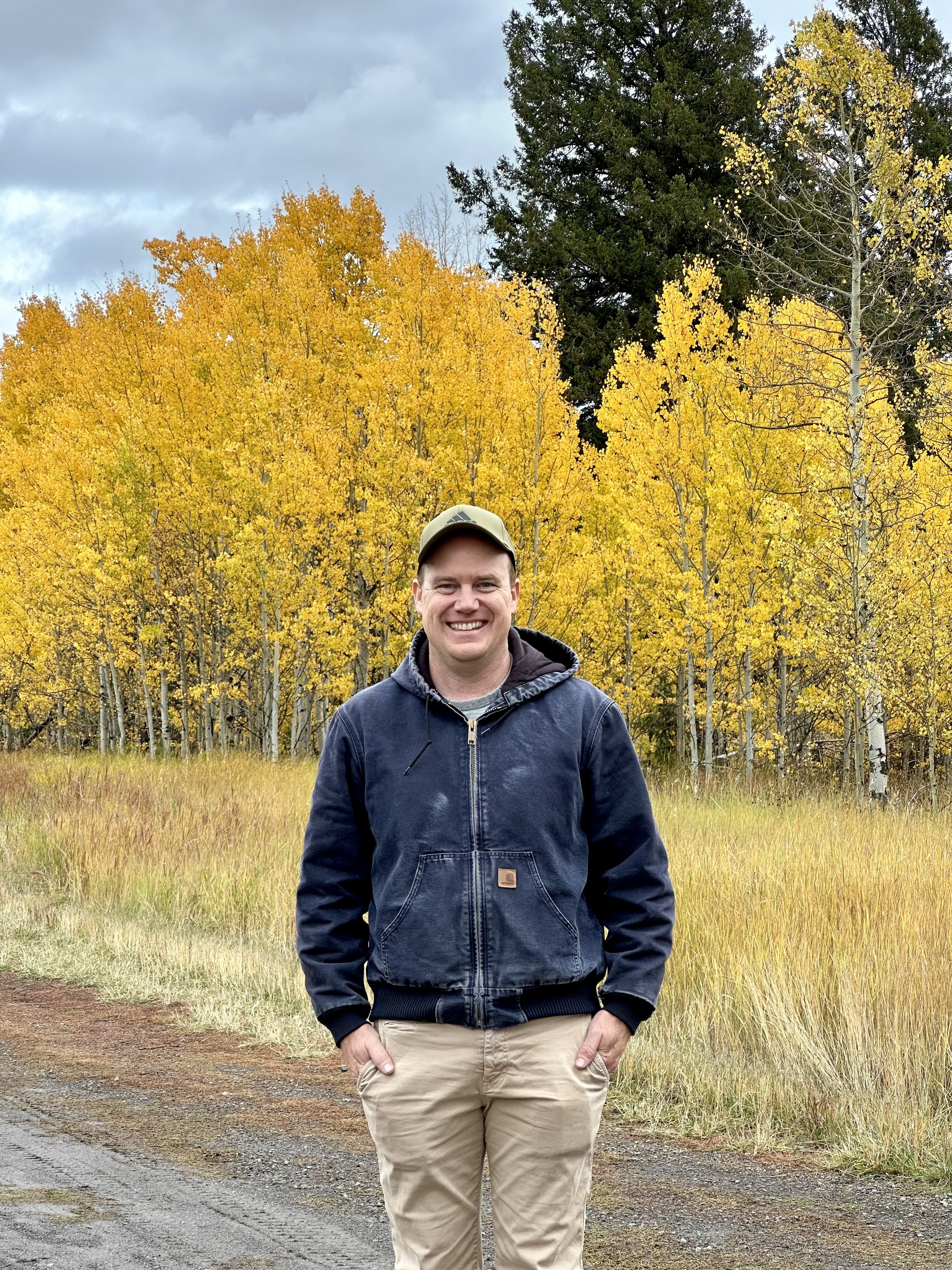 A smiling man in a dark blue jacket and khaki pants standing outdoors on a dirt path with yellow fall foliage and trees in the background.