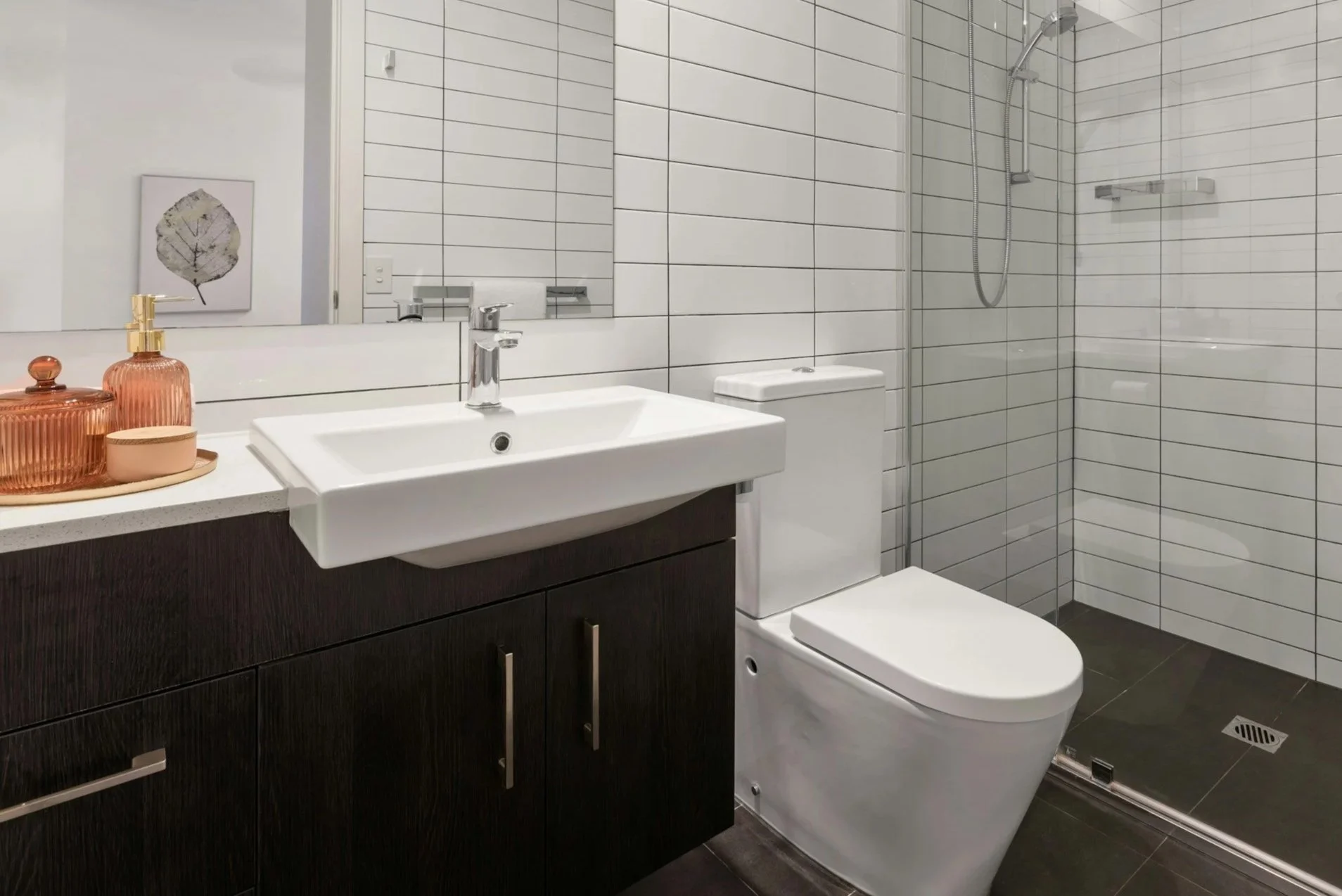 Modern bathroom with a white vanity sink, dark wood cabinet, toilet, and a walk-in shower with white tiles and a rain shower head.
