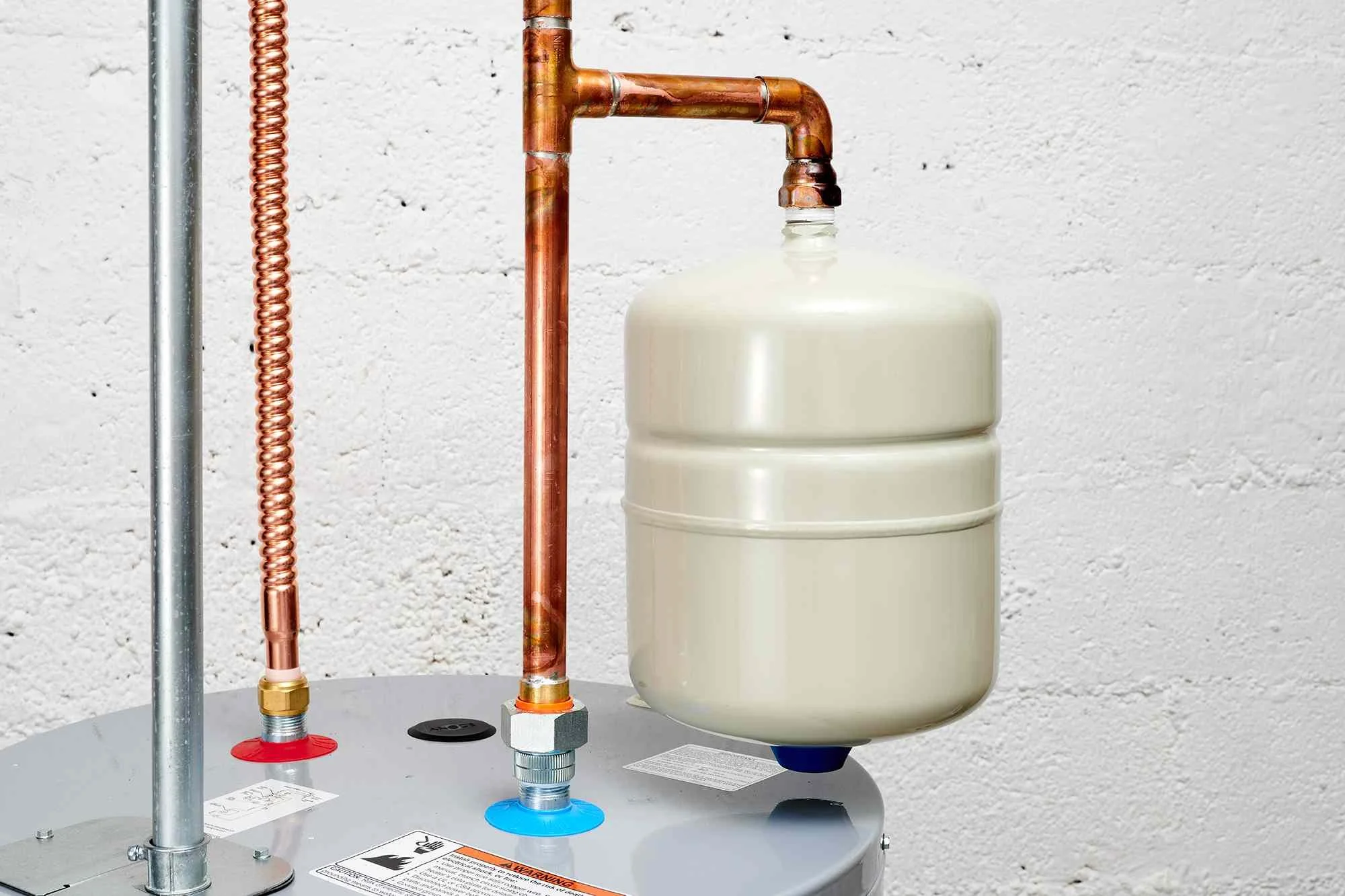 Close-up of a water heater with copper pipes and a white expansion tank against a white textured wall.