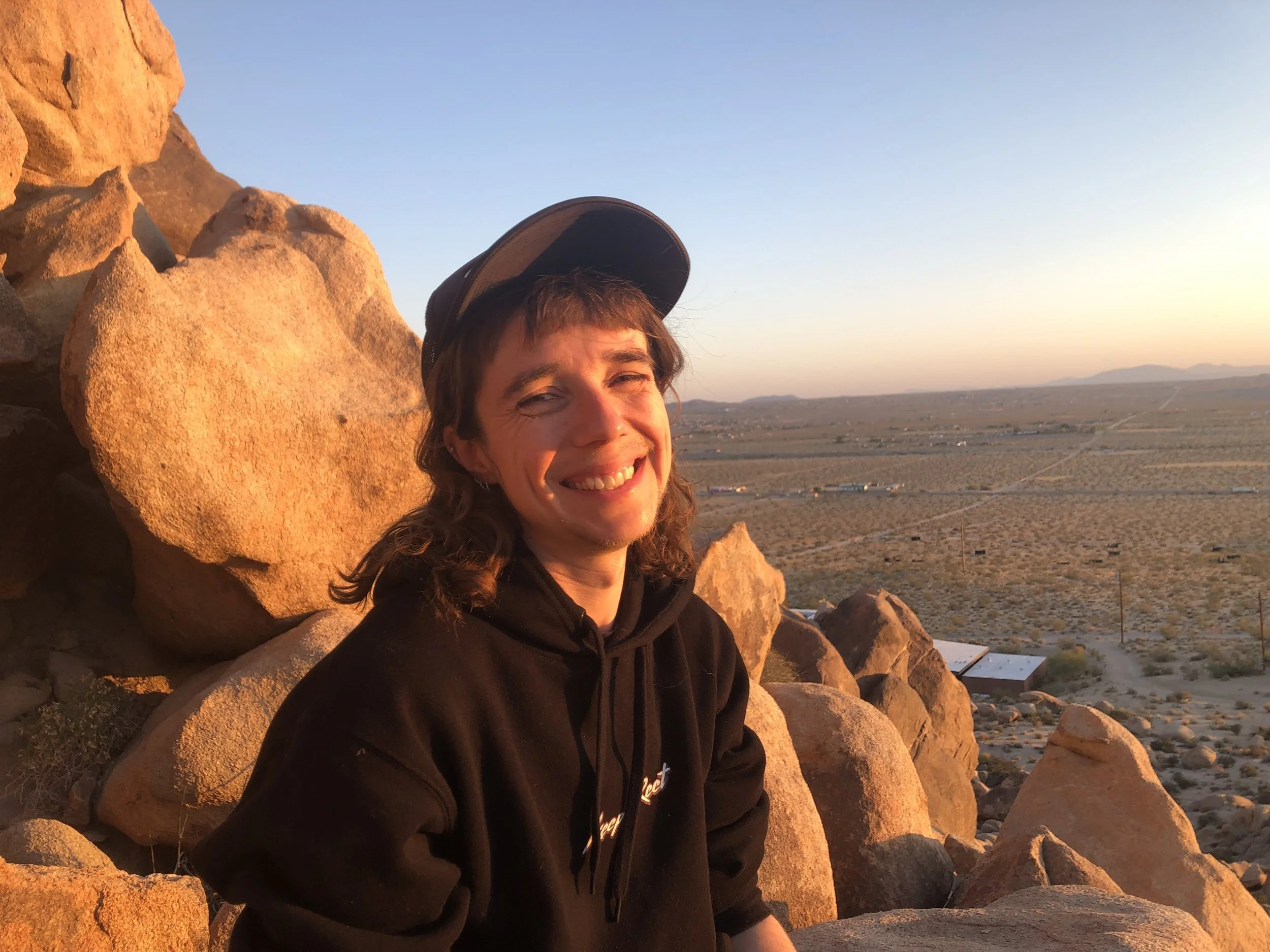queer person sitting smiling at a rocky lookout in front of a sunset