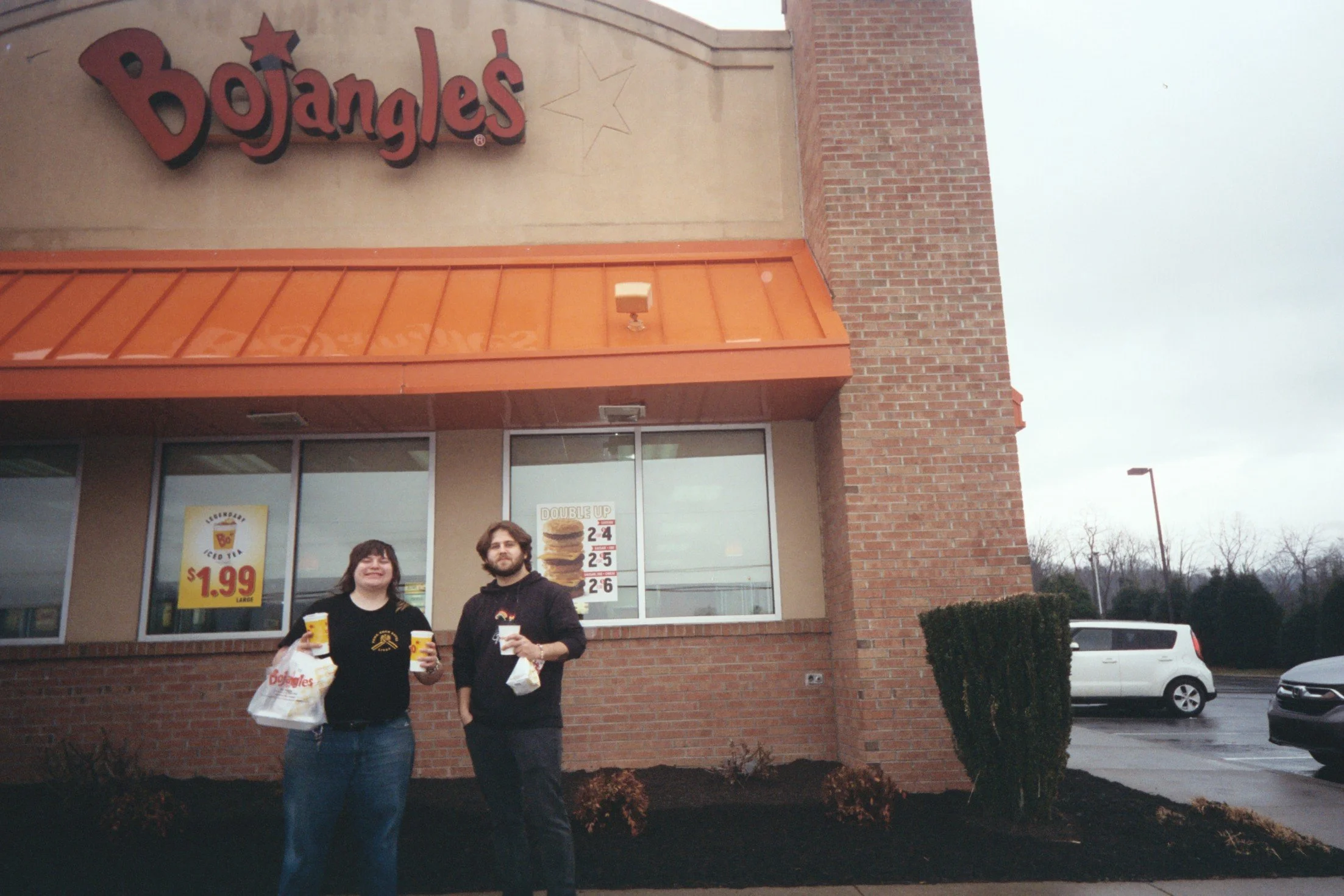 Frog Legs (Nano and Jacob) standing in front of a Bojangles with some takeout food, smiling.