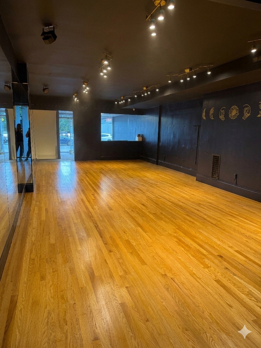 Empty dance studio with wooden floors, black walls, and ceiling lights, with a large mirror on the left wall and a window at the back.