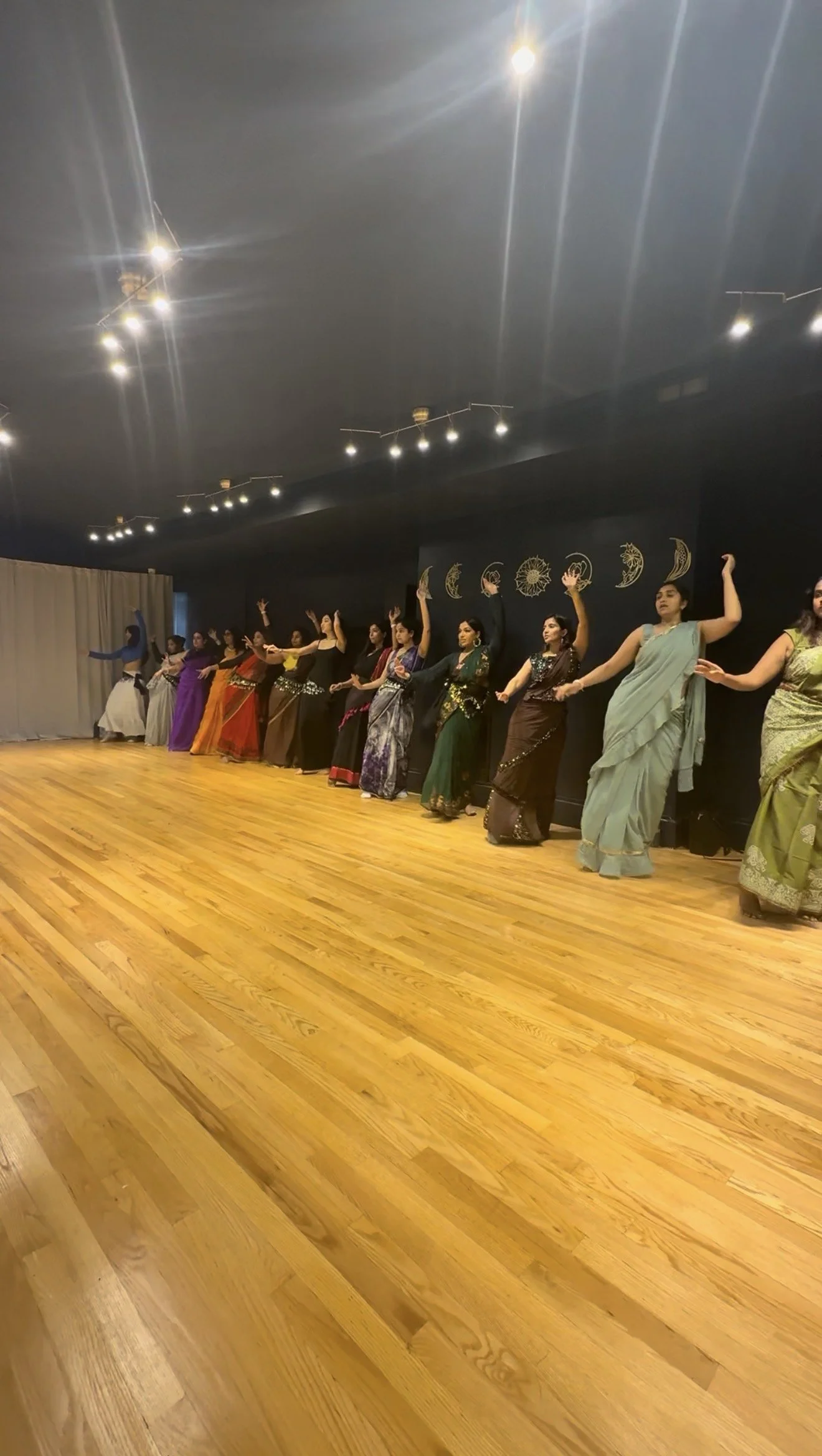 Group of women in colorful sarees performing a dance on wooden floor in a room with dark walls decorated with moon phases and celestial designs.