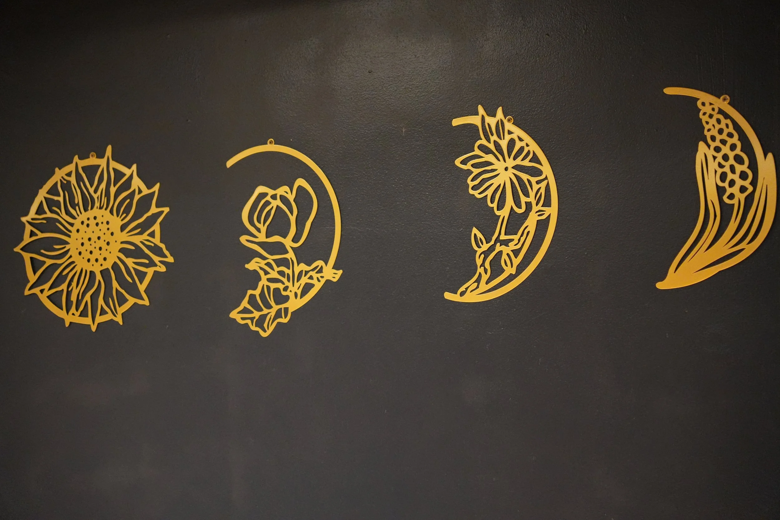 Four gold-colored cutting boards with floral and botanical cutout designs hanging on a dark wall.