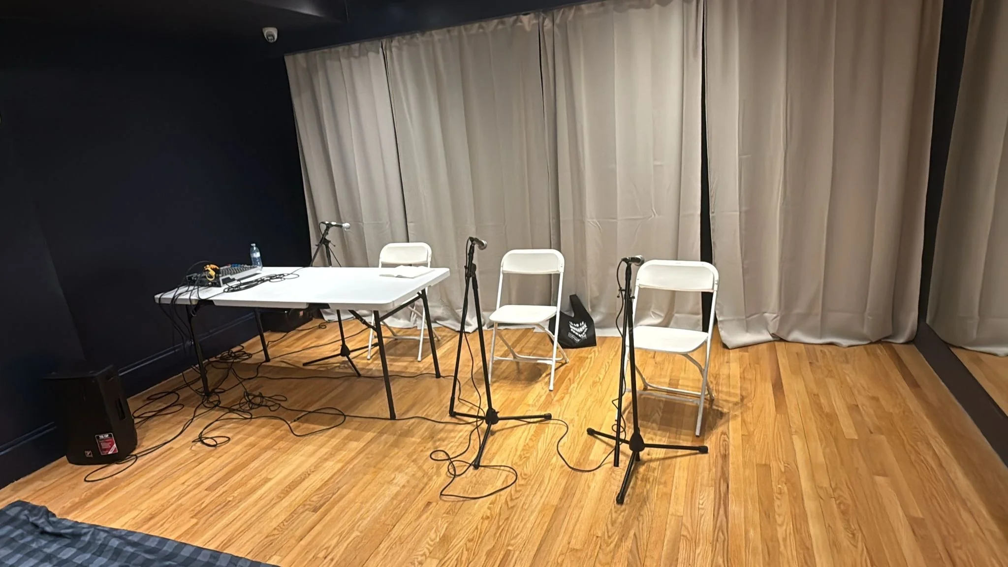 A small stage setup with three white chairs in front of a curtain, three microphones on stands, a table with audio equipment, and a bottle of water, on a wooden floor.