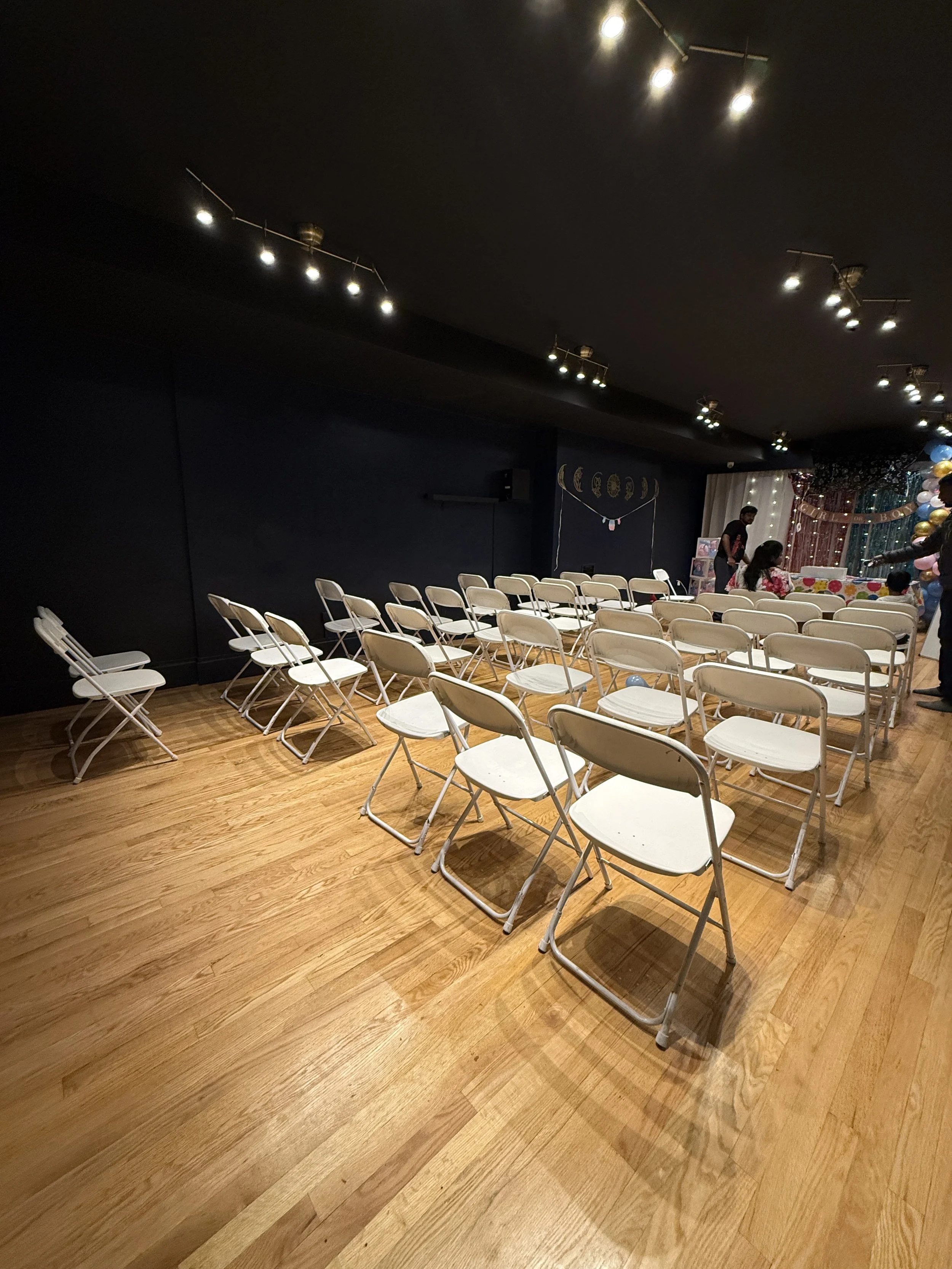 Empty white chairs arranged facing a decorated stage with balloons and people setting up for an event in a dark room with string lights.