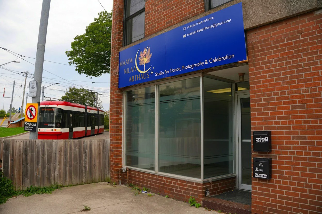 Exterior of Maaya Nilaa ArtHaus studio with a blue sign, brick walls, a glass window, and an entrance door. A streetcar is passing on the street nearby.