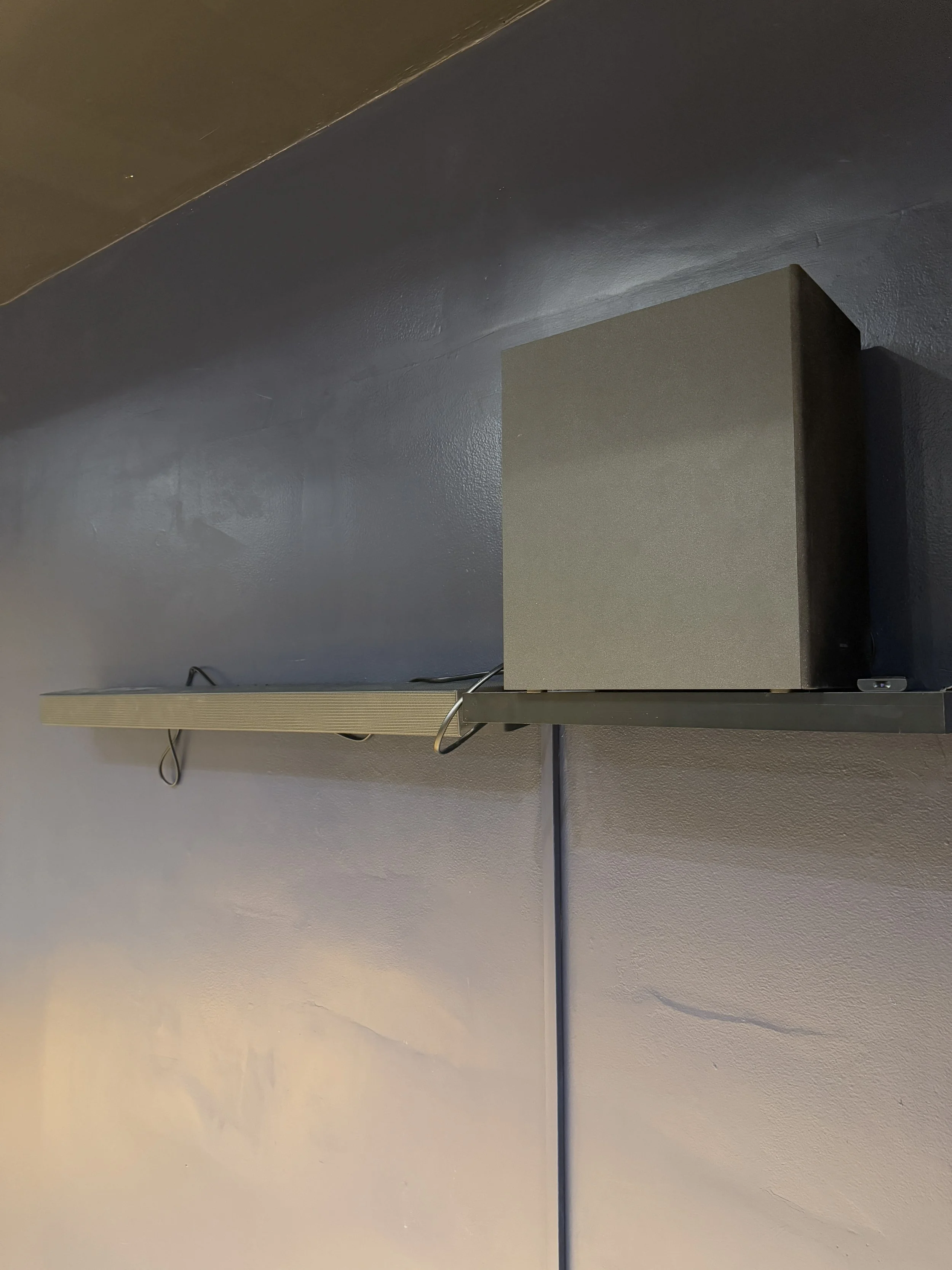 Wall-mounted black speaker on a metal shelf with electrical wires underneath, against a dark-colored wall.