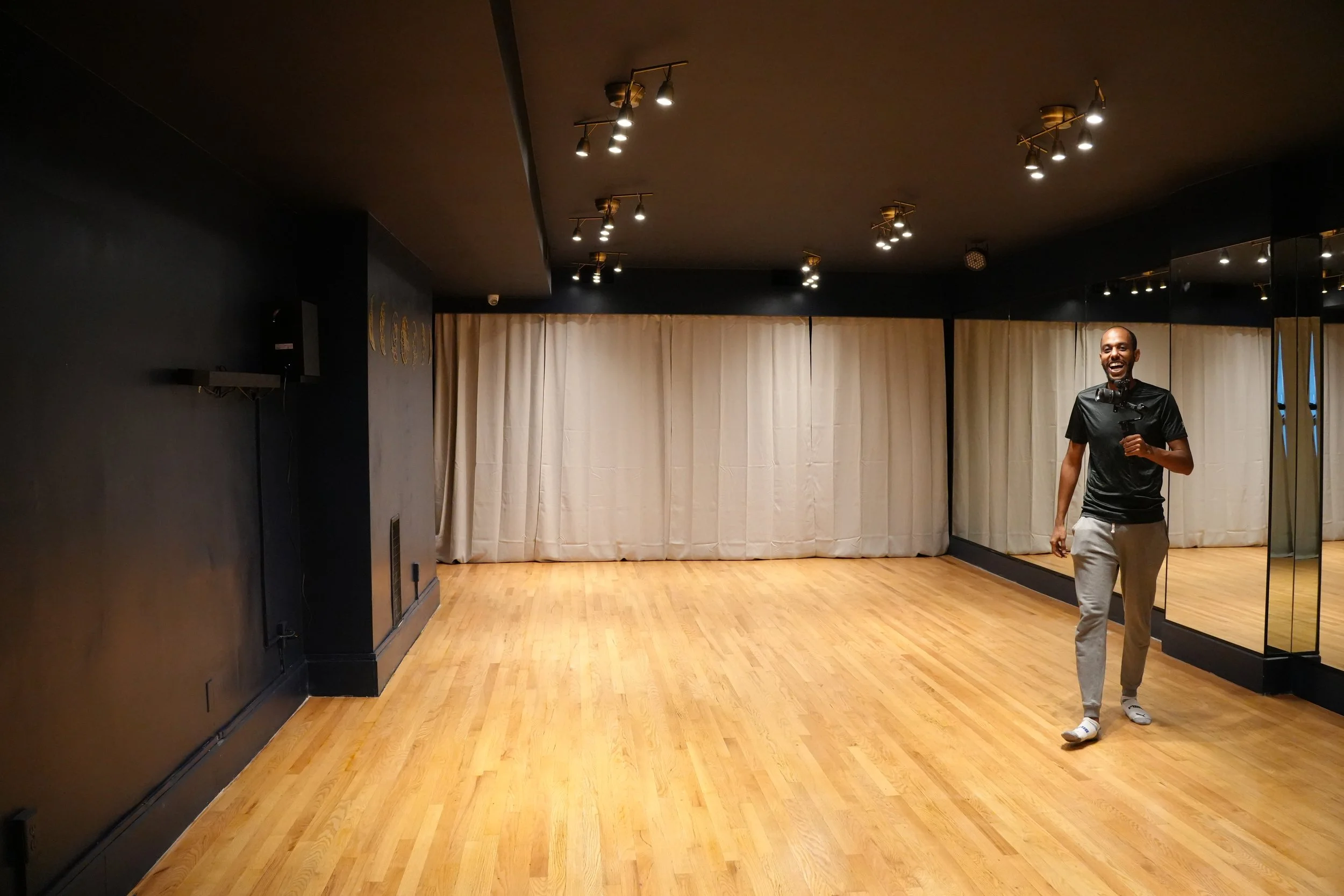 In a dance studio or rehearsal room with hardwood floors, black walls, and beige curtains, illuminated by ceiling spotlights.