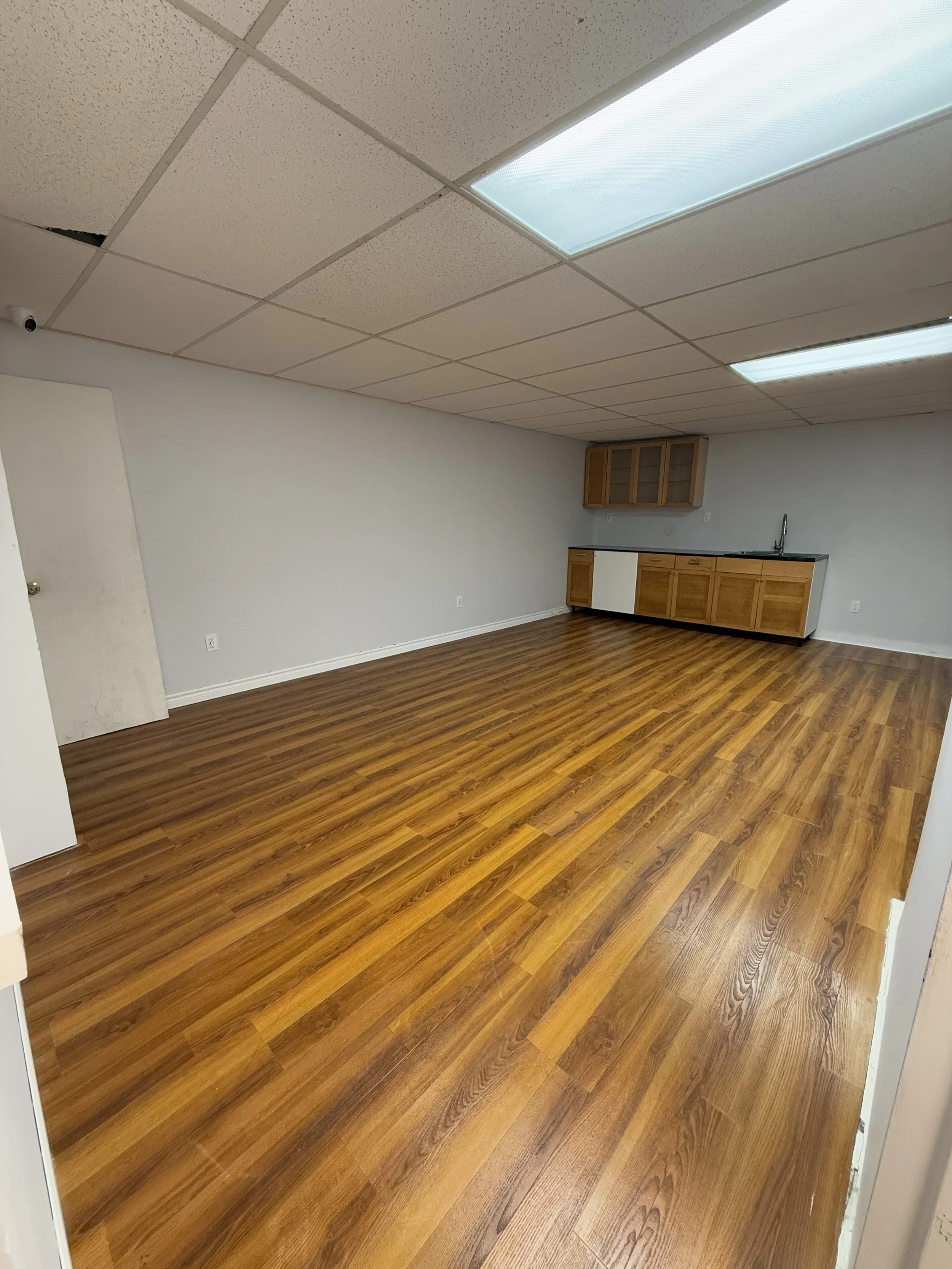 A large empty room with wood flooring, white walls, a drop ceiling with fluorescent lights, and a small kitchenette with upper and lower wooden cabinets, a sink, and a countertop.