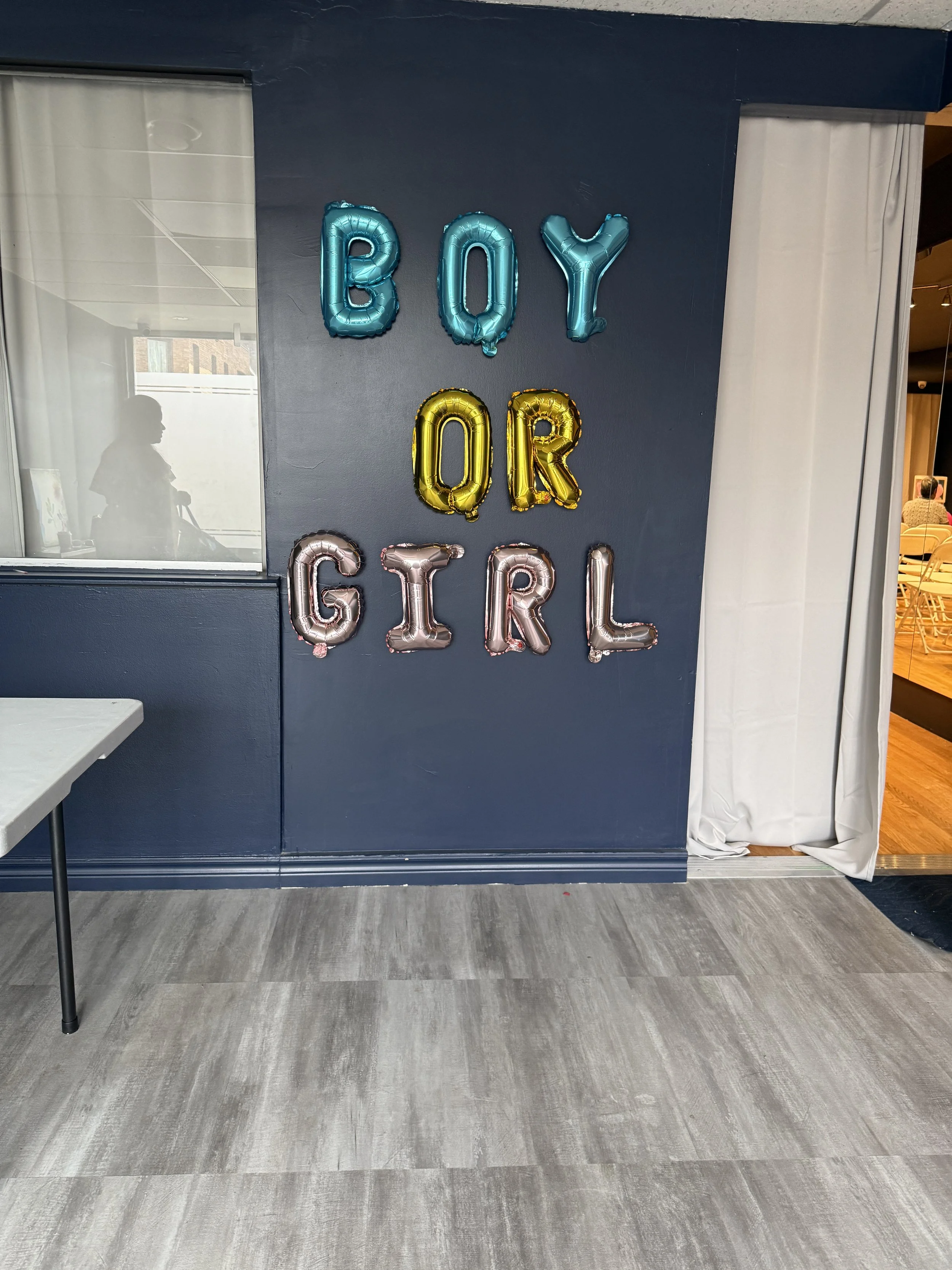 Decorative balloons spelling out "BOY OR GIRL" against a navy blue wall in a room, likely for a gender reveal celebration.