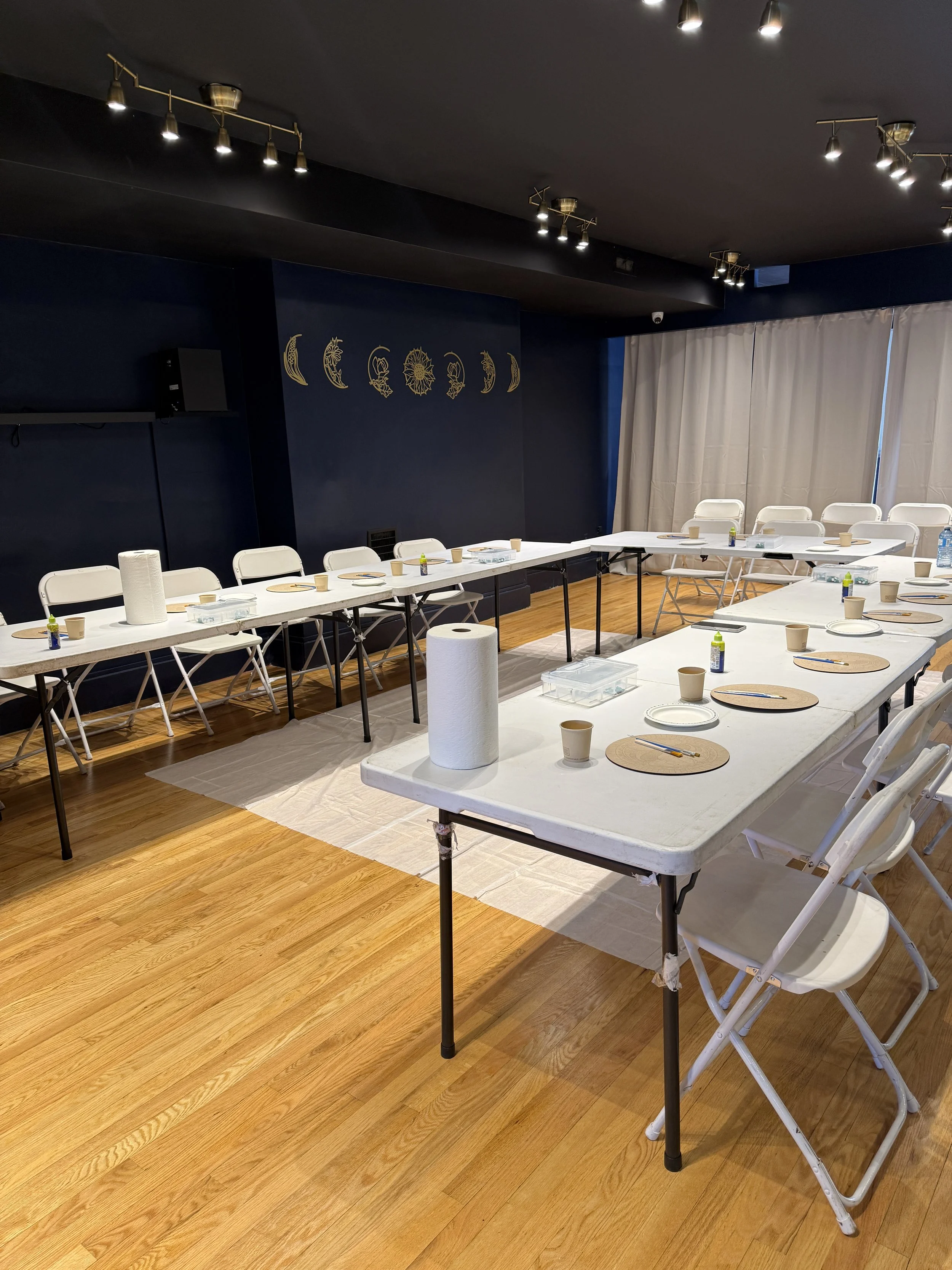 A room with white folding chairs and long tables set with paper plates, cups, and utensils, arranged for a gathering or event, with a dark blue wall decorated with moon and sun designs and bright overhead lighting.