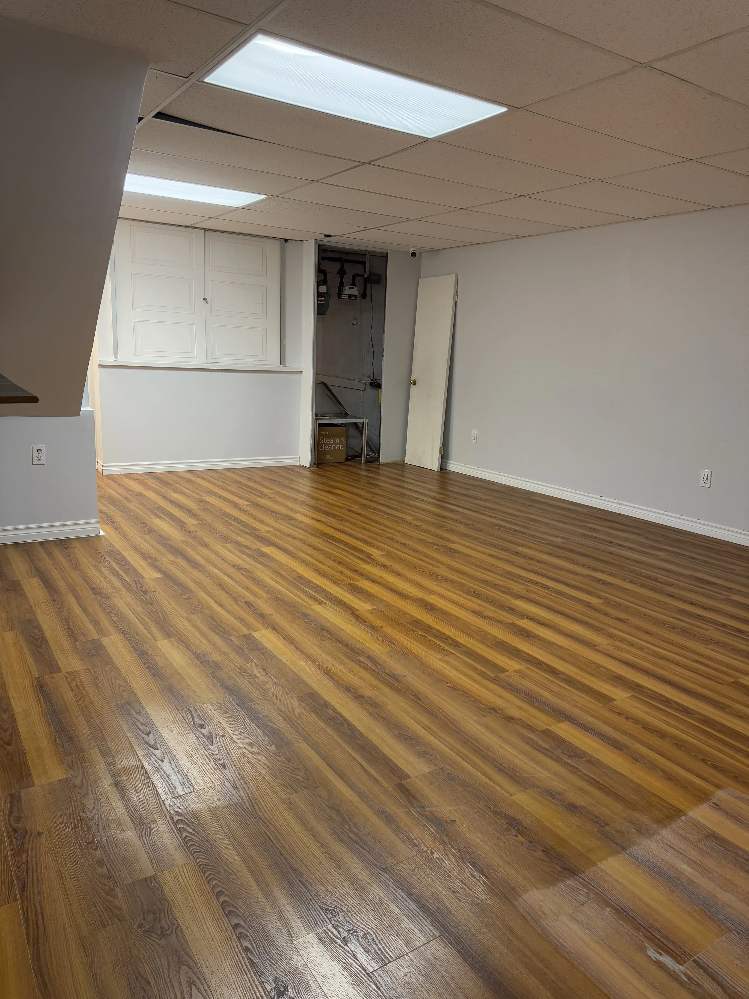 Empty basement room with wood flooring, white walls, white ceiling tiles, and a closed white door, with a utility area visible through an open doorway.