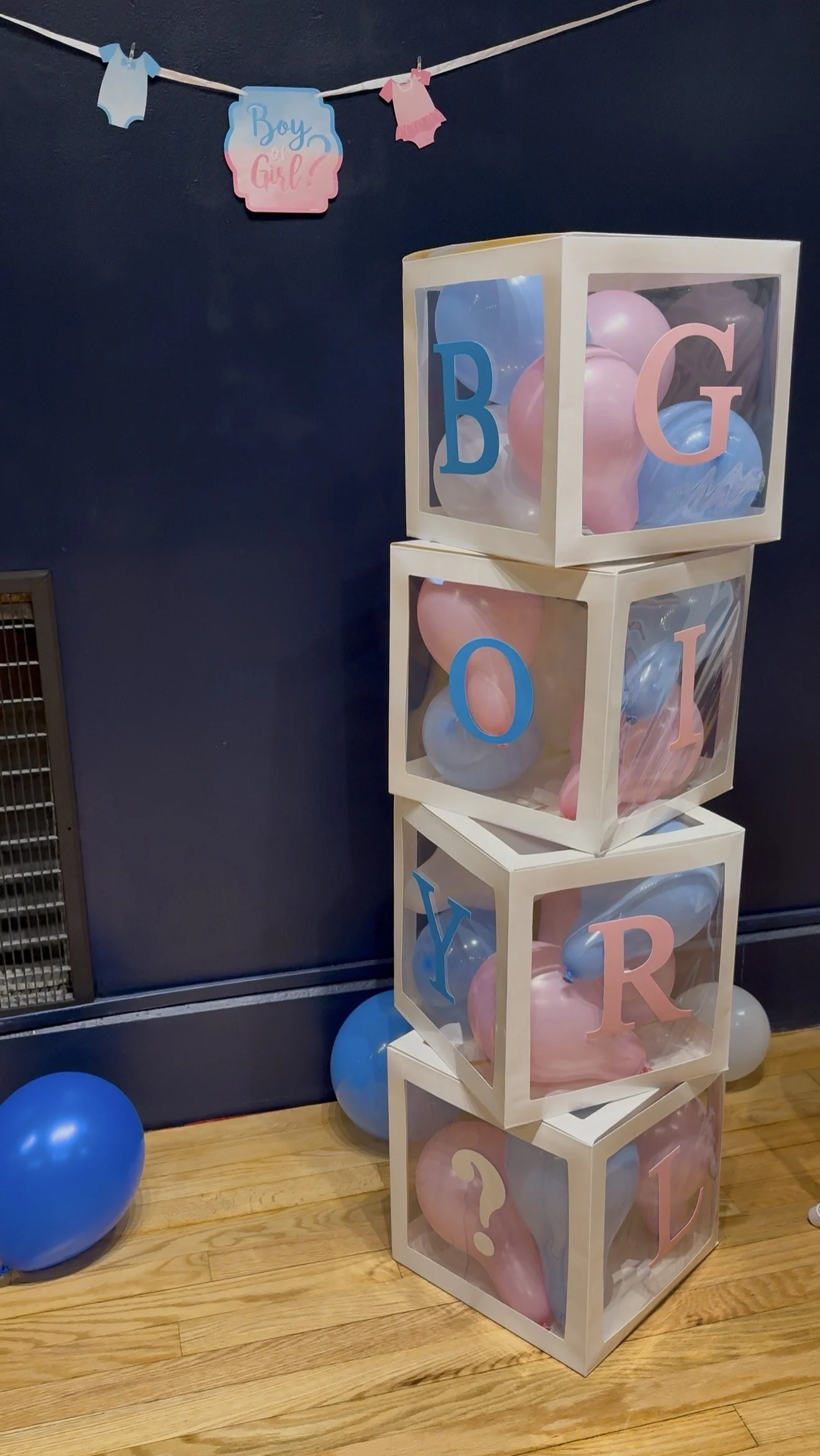 Four clear boxes stacked vertically, each filled with pink and blue balloons, with colored letters on the front spelling "BOY," "GIRL," "OPEN," "YARD," and a question mark. Blue and pink balloons are scattered on the wooden floor nearby, and a banner