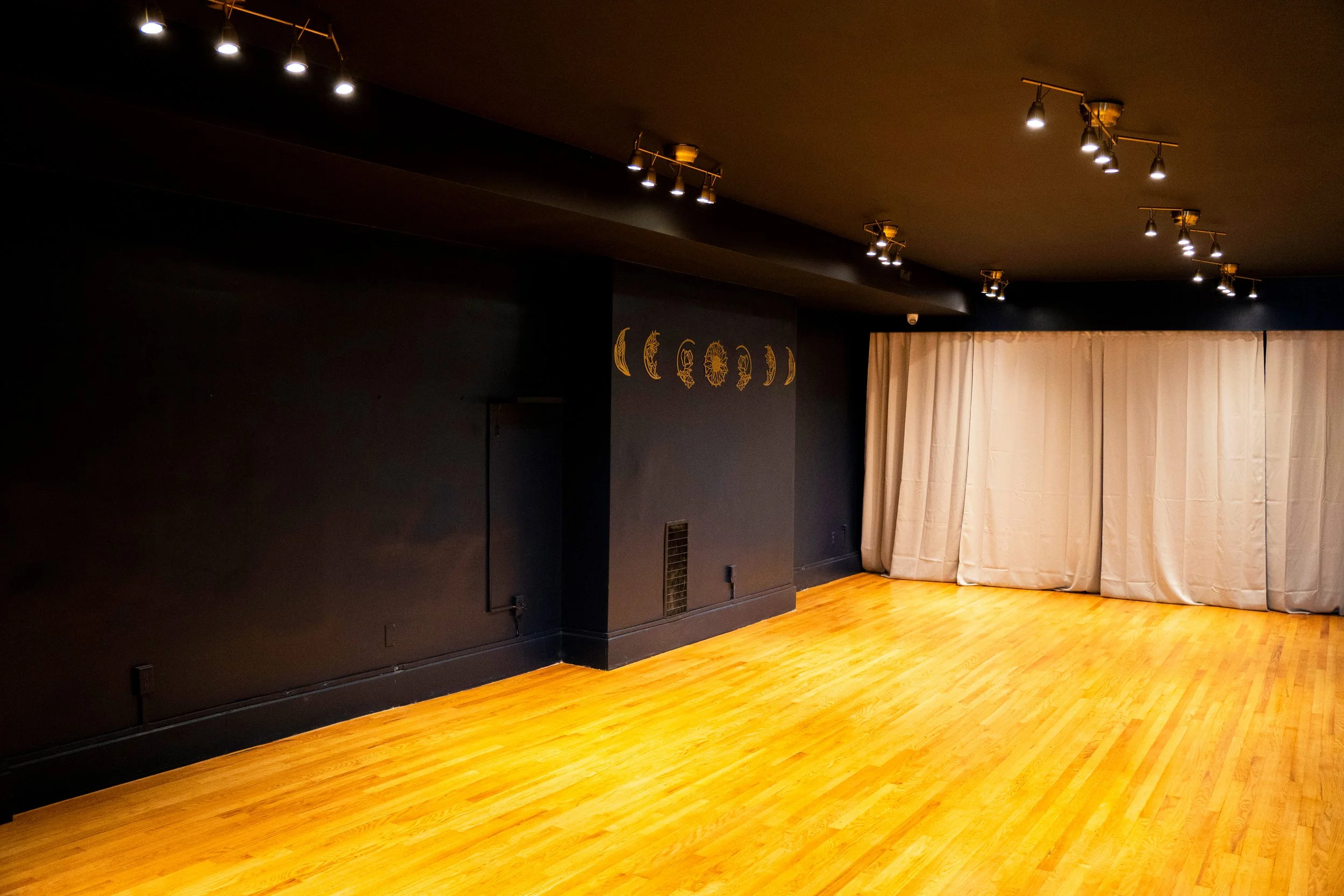 Empty performance or dance studio with wooden floor, black walls, and grey curtains at the back. Overhead lighting fixtures illuminate the space.