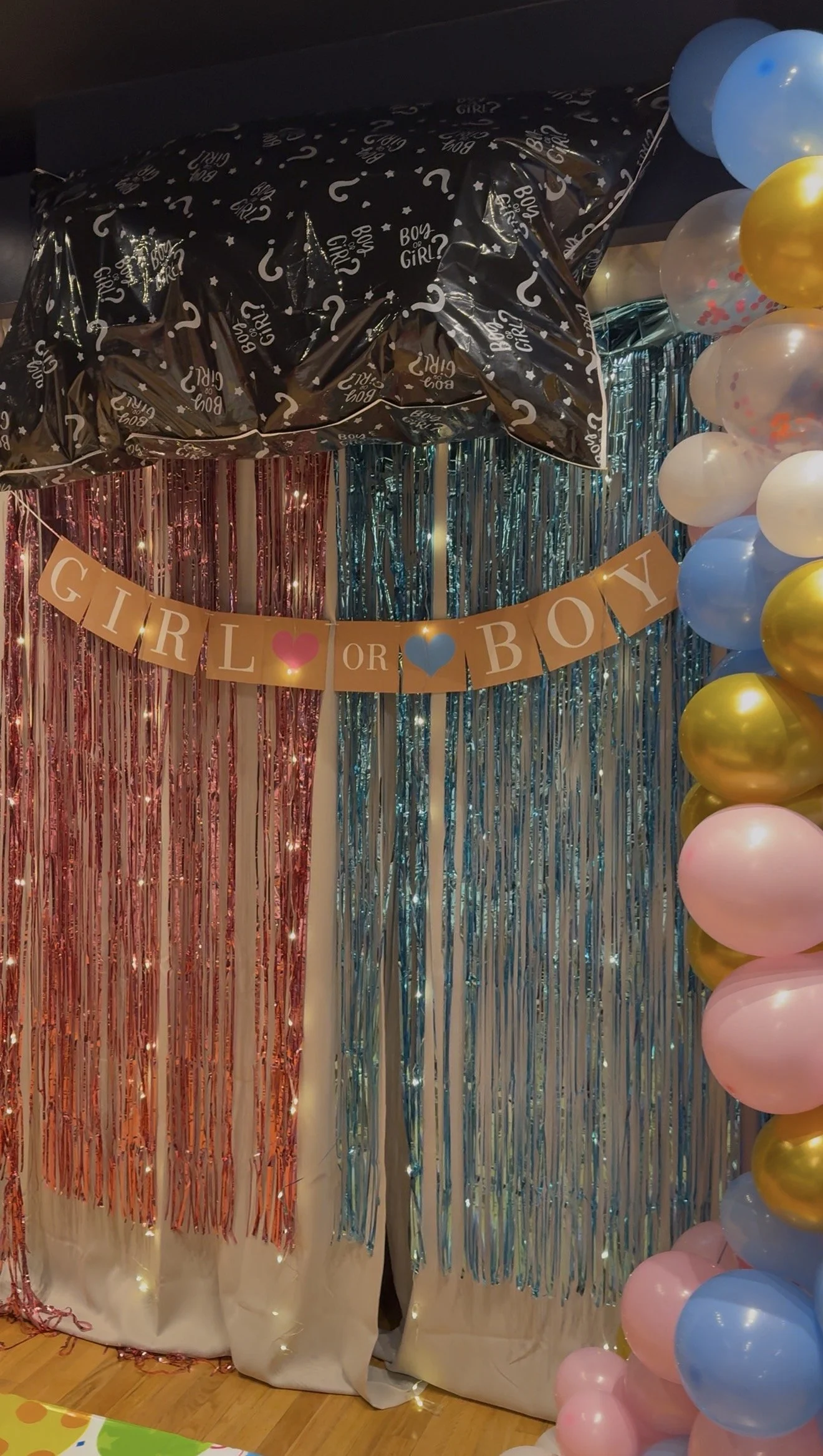 Decorative backdrop with metallic fringes in pink and blue, a banner reading 'Girl or Boy' with pink and blue hearts, and an arch of pastel-colored balloons including pink, blue, white, and gold.