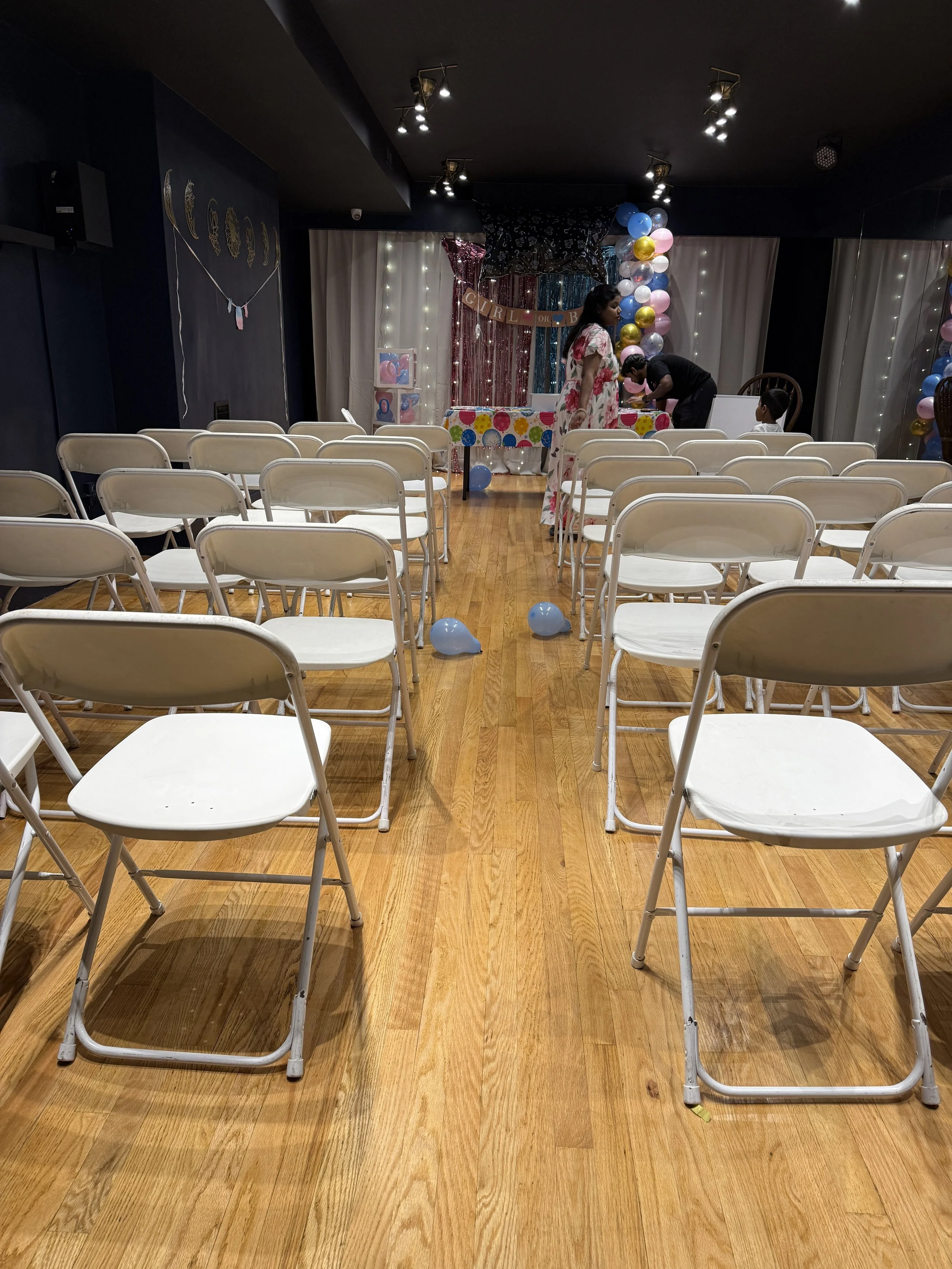 Decorated room with white chairs arranged in rows, balloons, and a table at the front, indicating a celebration or party setup for gender reveal, baby shower or birthday party