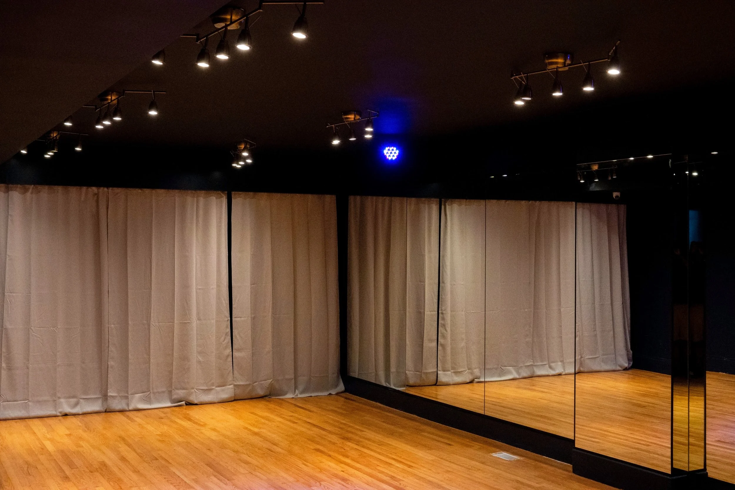Dance studio with beige curtains, wooden floor, black walls, mirrored wall, and ceiling track lighting with spotlights.
