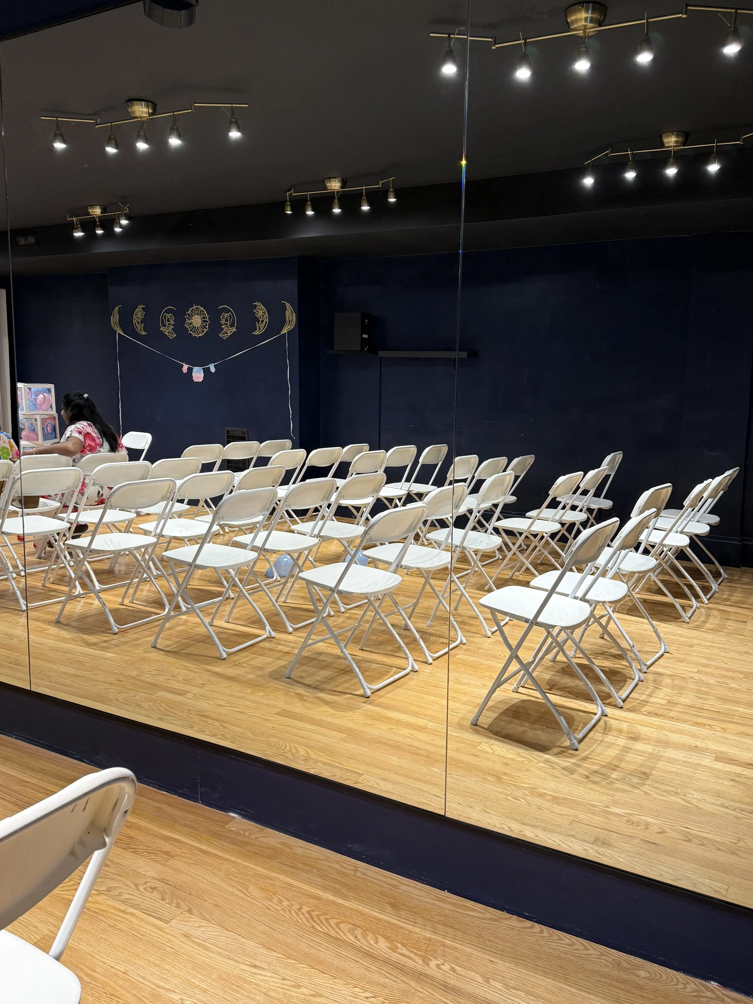 Empty white folding chairs arranged in rows facing a stage with decorations of moon phases and hanging ornaments, in a room with wooden floor and dark blue walls, reflected in a glass wall. arty setup for gender reveal, baby shower or birthday party
