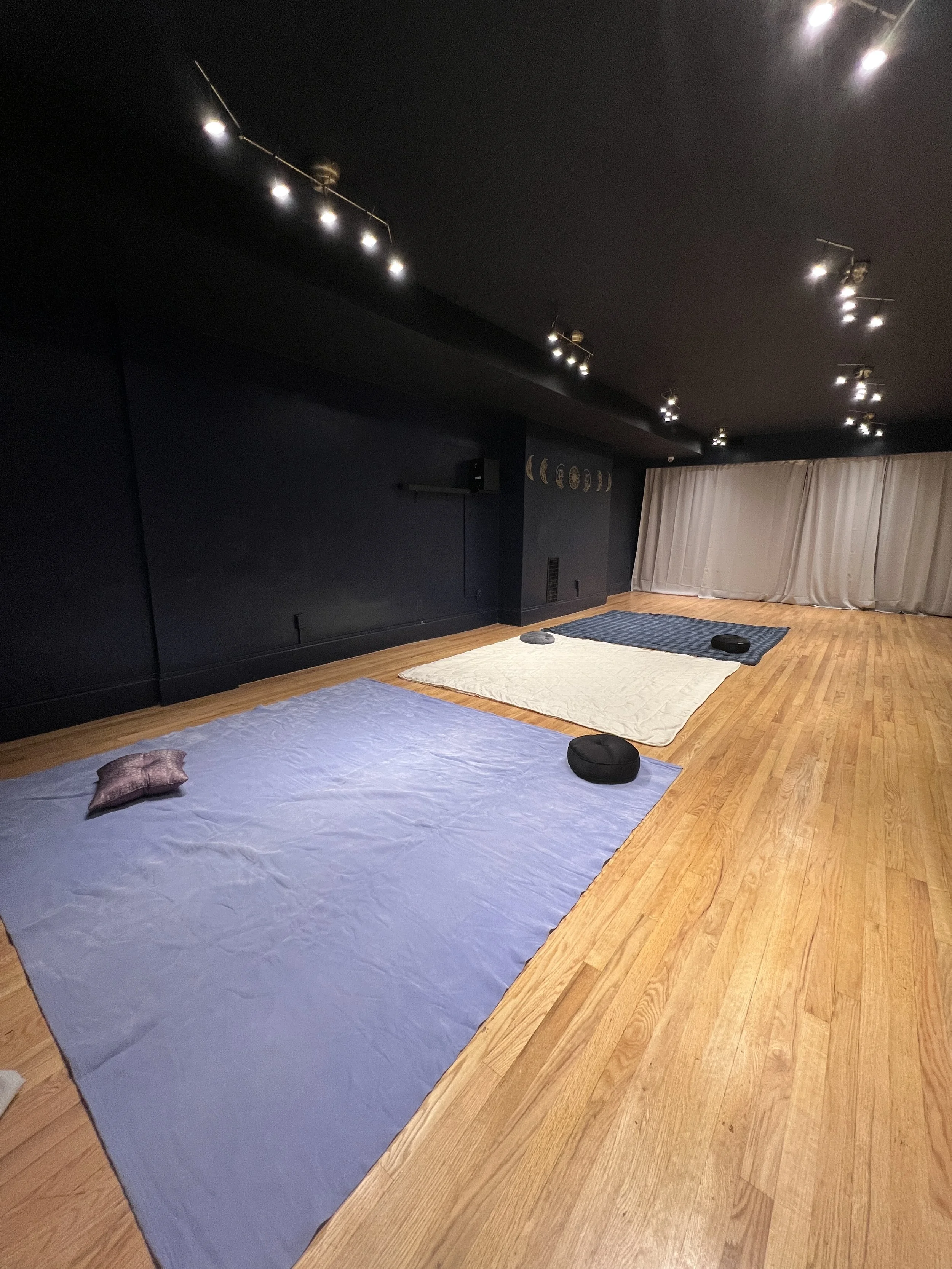 A room with hardwood floors and dark walls, set up with three large mats and cushions on the floor, possibly for a meditation or yoga session.