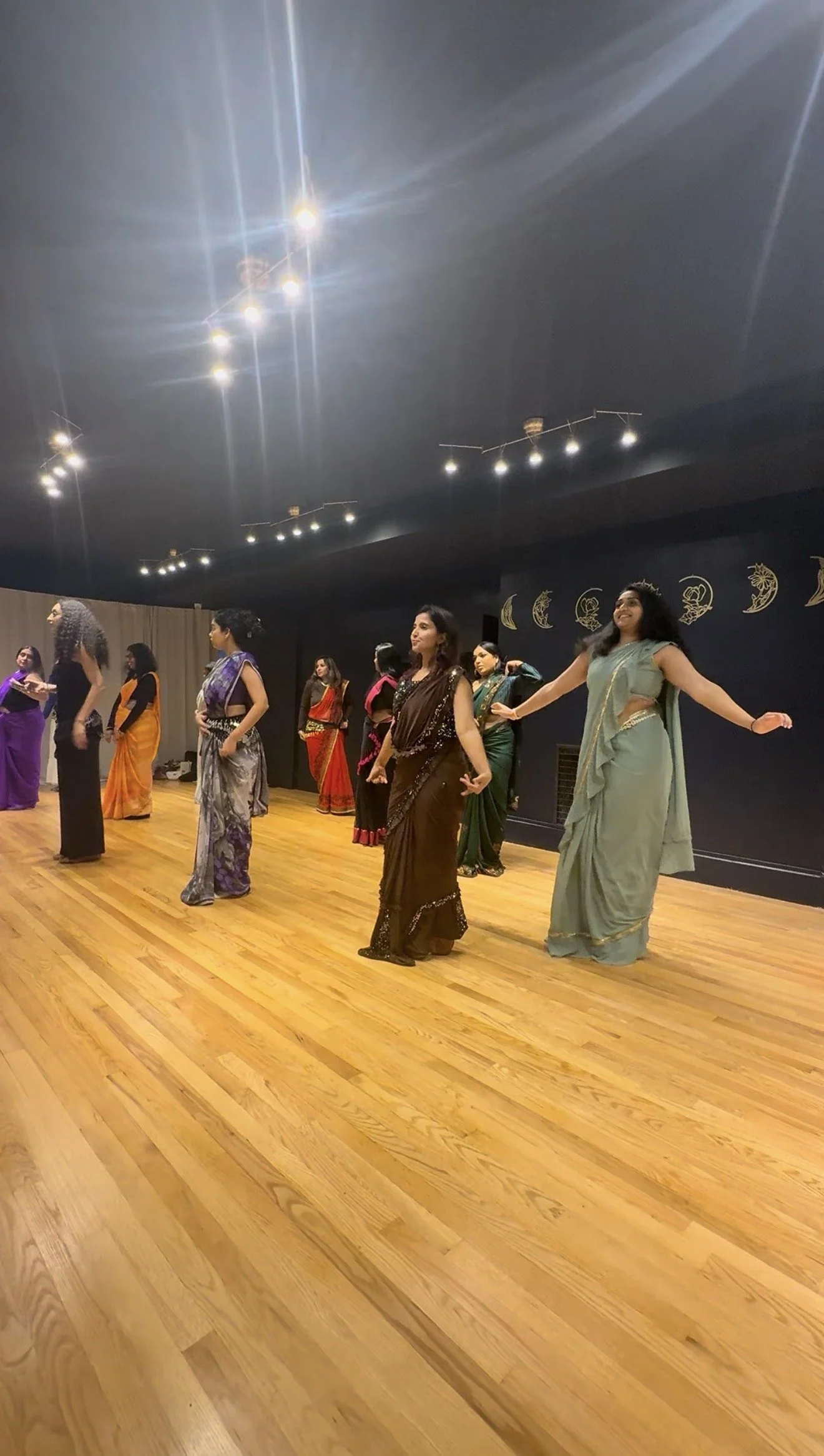 A group of women in colorful sarees performing a dance or yoga routine in a room with wooden floors and a dark ceiling decorated with moon and star designs.