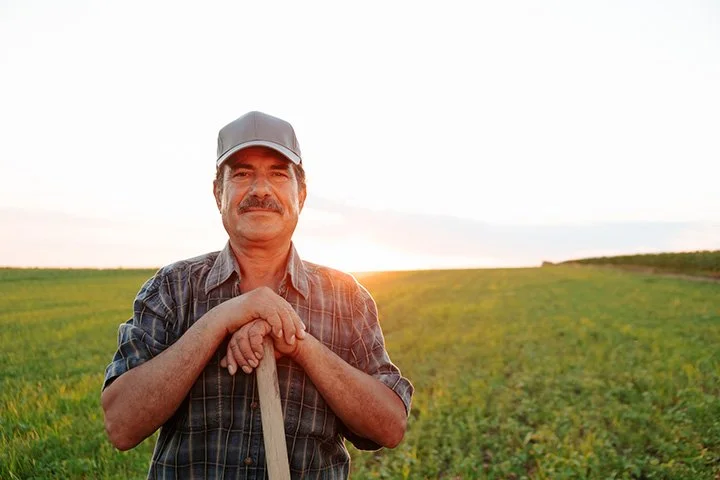 A middle-aged man with a mustache, wearing a plaid shirt and a gray cap, standing outdoors in a green field during sunset, holding a garden rake.