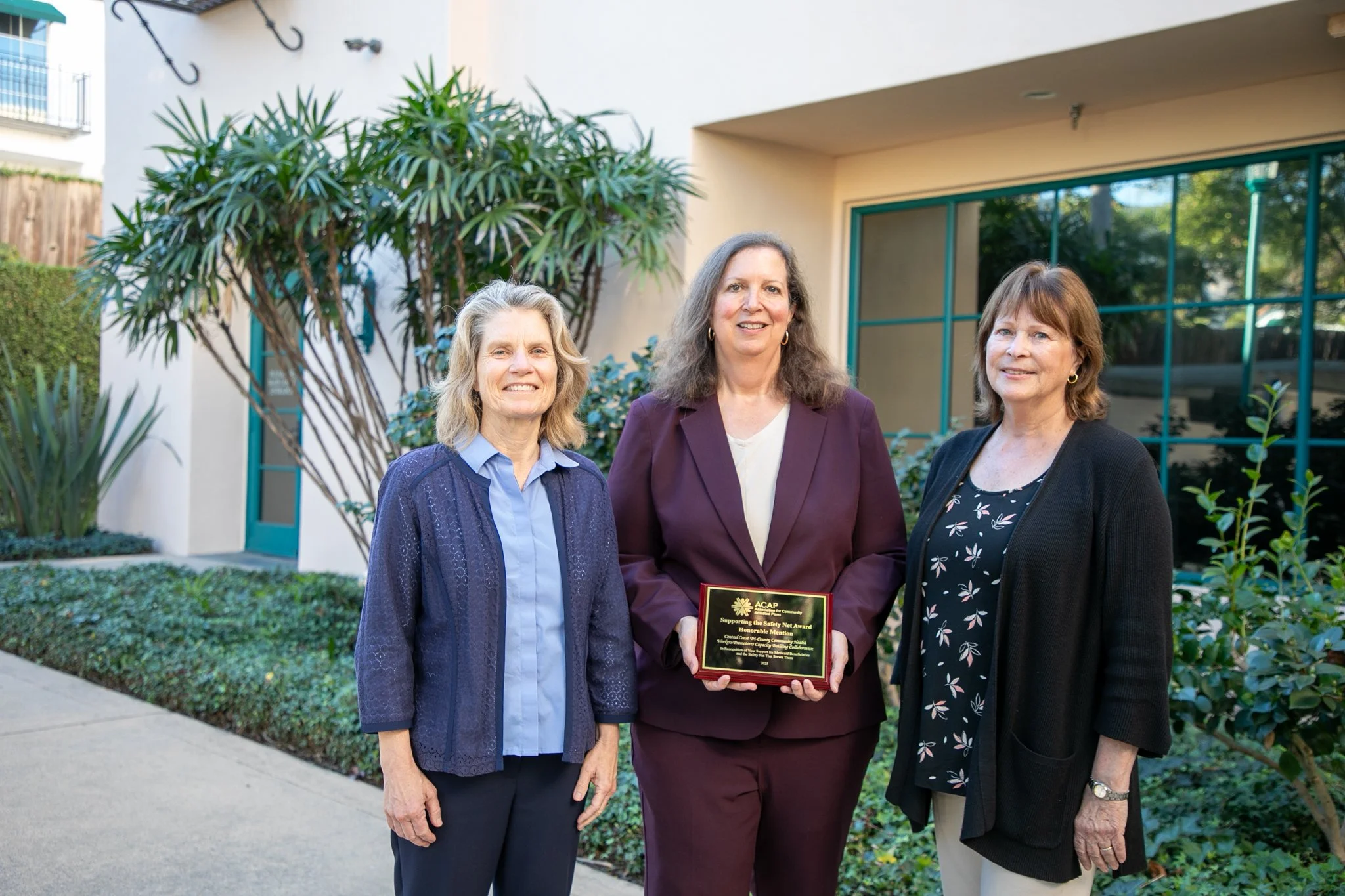 Three women standing outside in front of a building with large windows and greenery. The woman in the middle is holding a plaque. All are smiling.