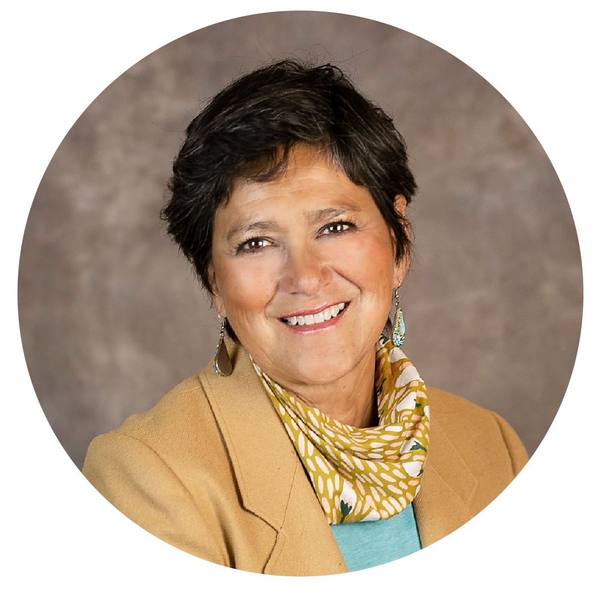 Headshot of a smiling middle-aged woman with short dark hair, wearing a tan blazer, yellow and white patterned scarf, blue top, and earrings, against a gray background.