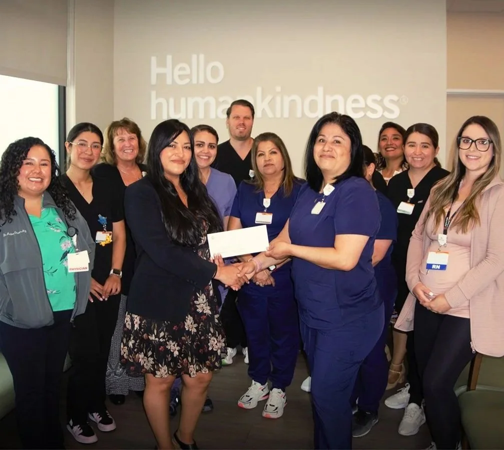 Group of healthcare professionals, including nurses and a physician, standing in front of a wall with the words 'Hello human kindness.' The group is smiling and one woman is receiving a donation or award.