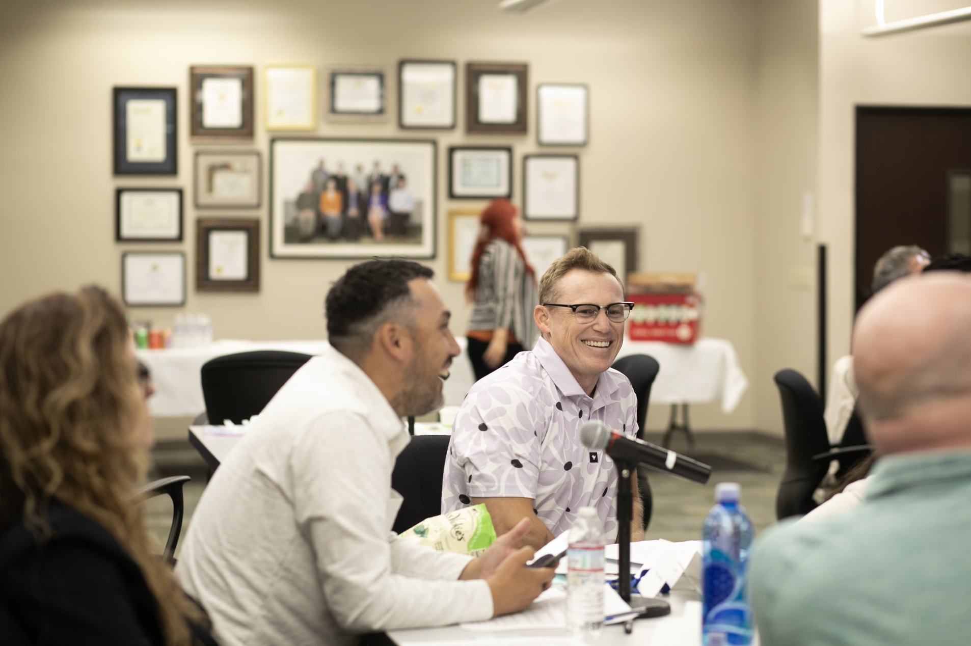 Group of people sitting at a table, with a man in glasses smiling and speaking into a microphone.