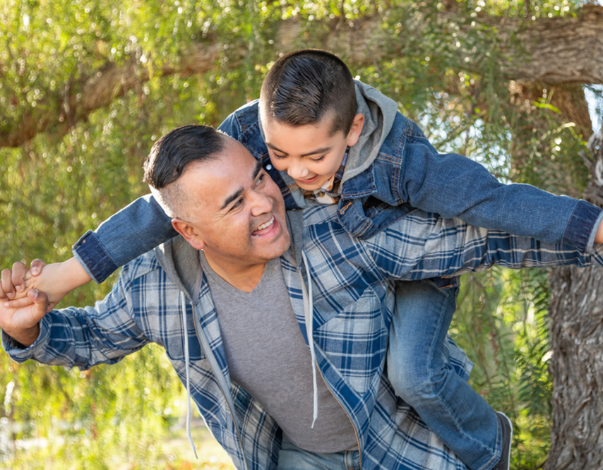 Father and son playing outdoors in a wooded area, with the father carrying the son on his back, both smiling and enjoying themselves.