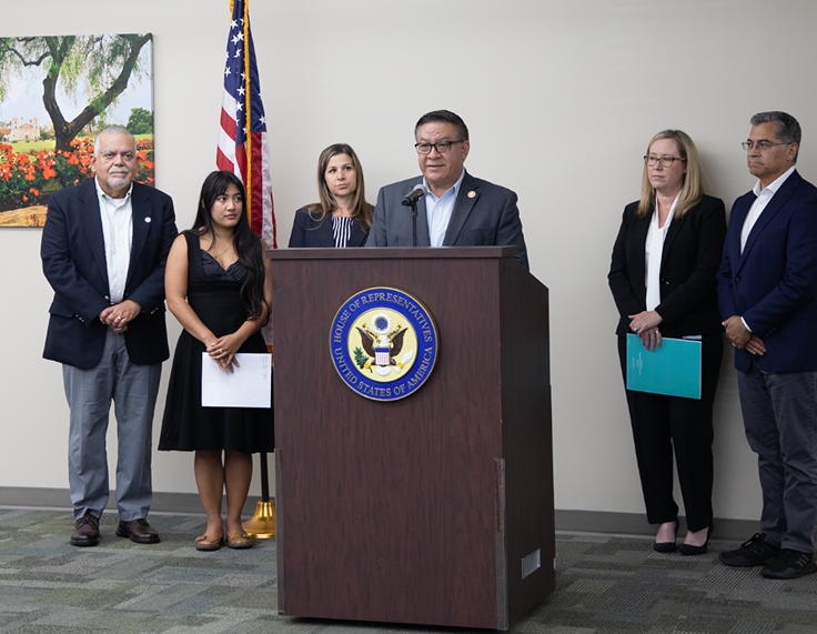 A group of six people standing behind a speaker at a podium with the House of Representatives emblem, in a room with an American flag and a landscape painting in the background.