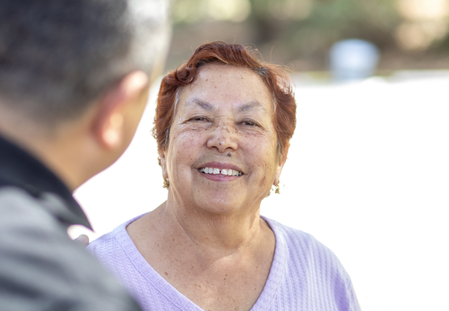 An elderly woman with short red hair smiling outdoors, talking to a man. She wears a lavender top and earrings.