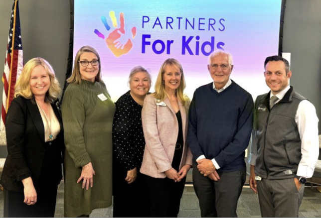 Six smiling people standing side by side in front of a screen displaying the logo 'Partners For Kids' with colorful handprints. The group includes five women and one man, all dressed in business casual attire, with an American flag partially visible 