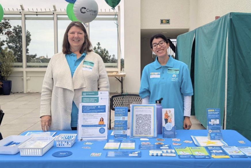 Two women standing behind an informational table at an outdoor event, smiling, with balloons and brochures on the table.