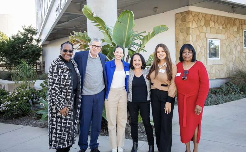 Six diverse women standing outdoors in front of tropical plants and a building, smiling for a group photo.