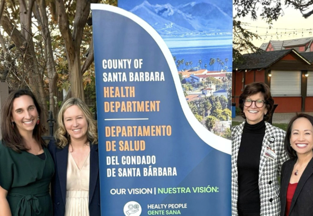 Four women standing outdoors next to a banner for the County of Santa Barbara Health Department, with trees and a building in the background.