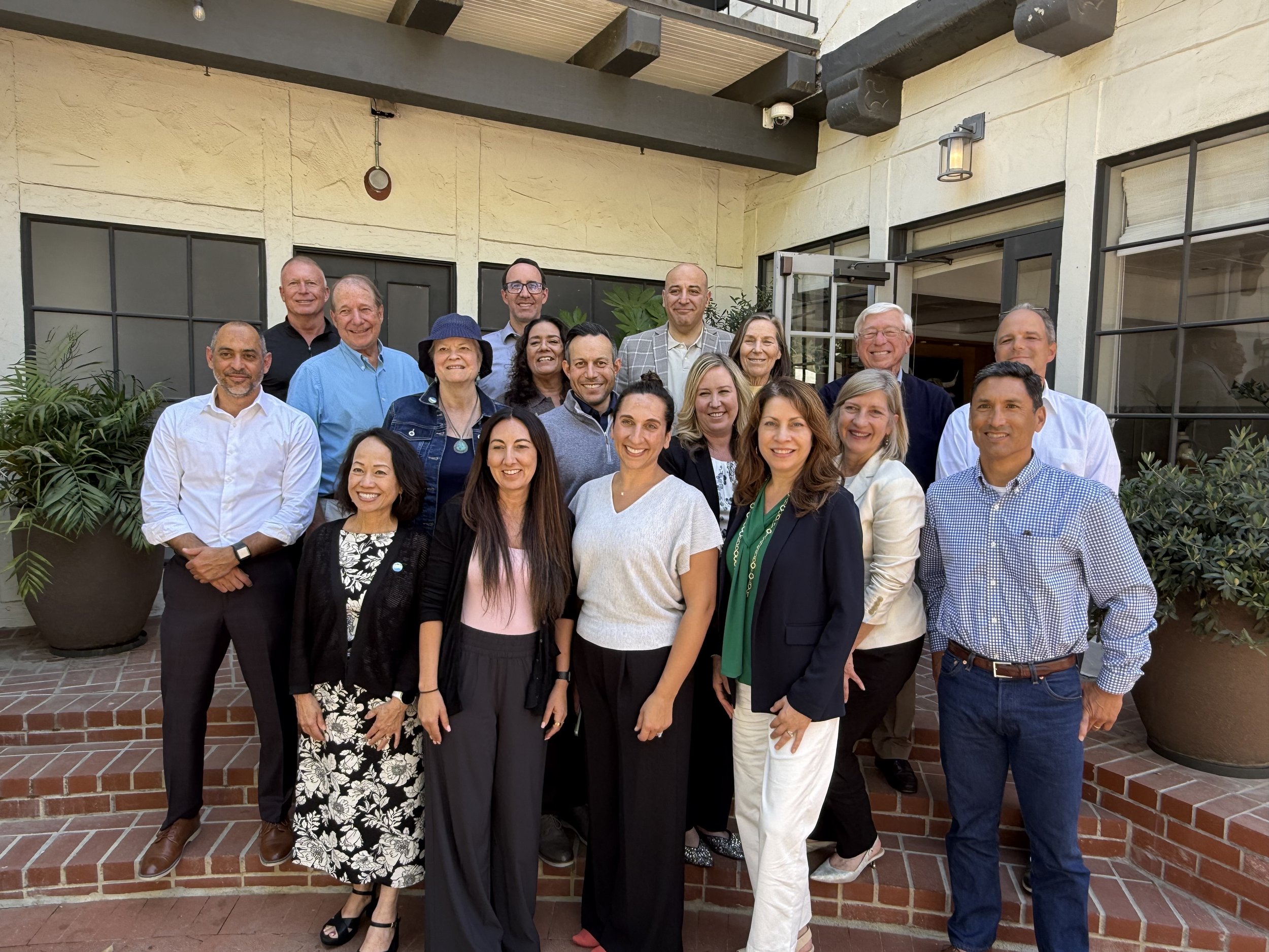 A group of 17 diverse adults posing for a photo on a set of brick stairs outside a building with large windows and potted plants.